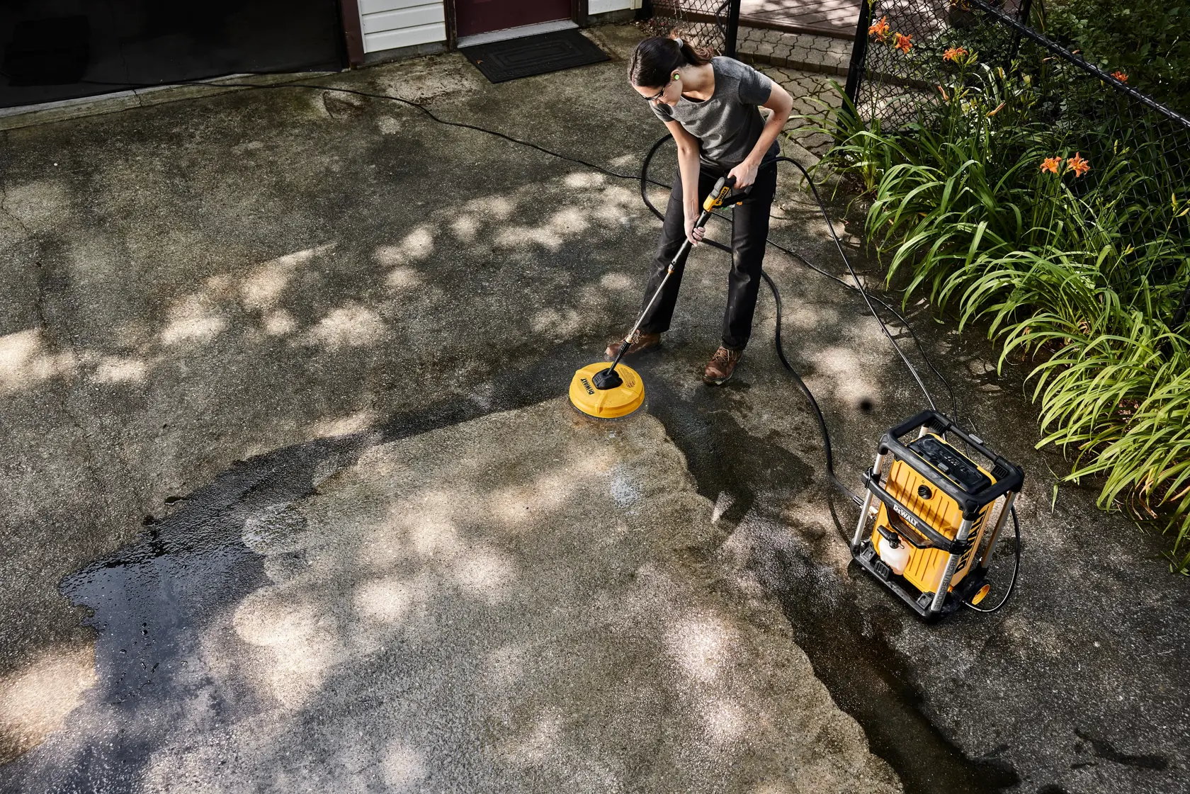 A person using a DEWALT DWPW3000 pressure washer to clean a concrete driveway, with the machine and cleaning attachment visible. The driveway is partially cleaned and there are plants along the side.