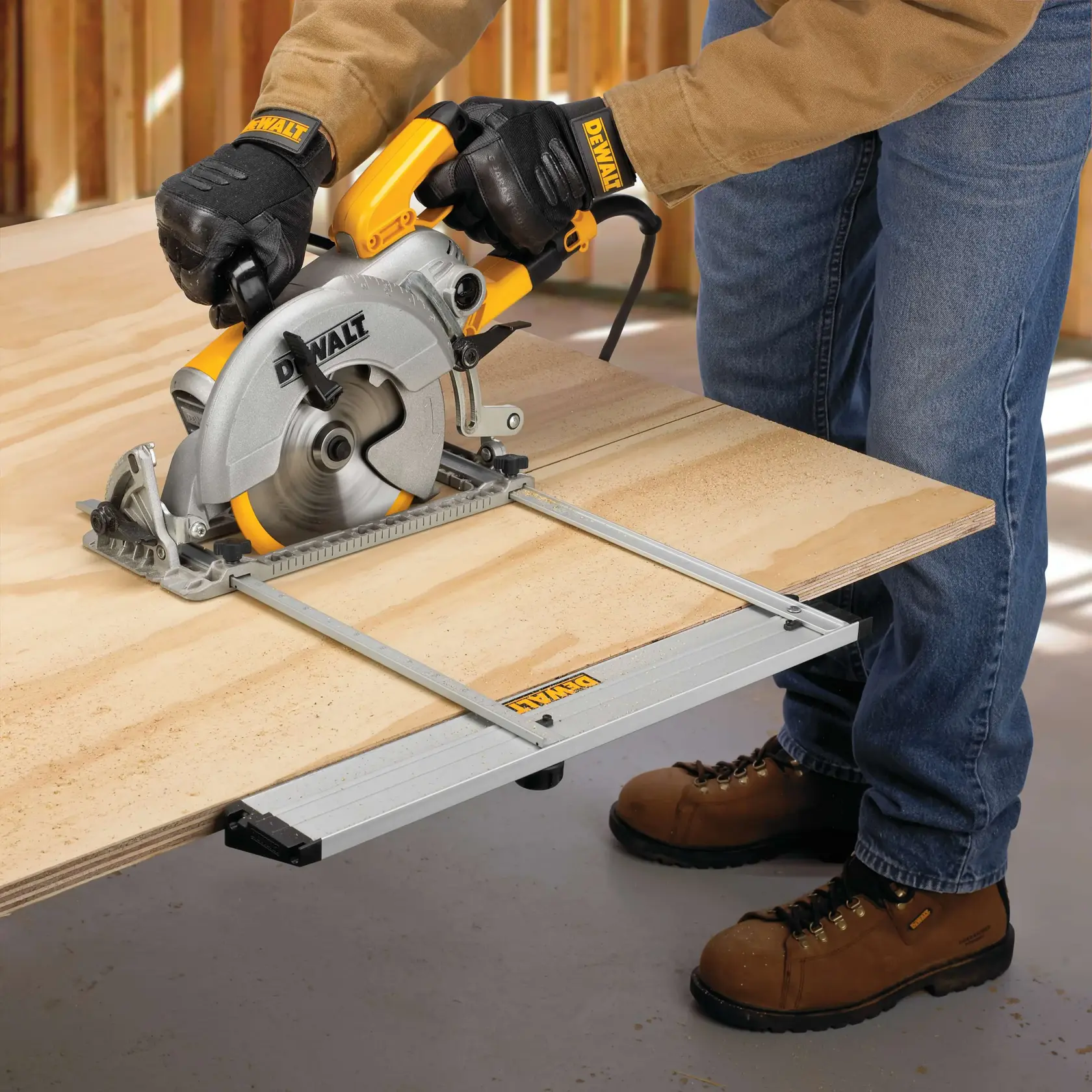 A person wearing gloves and work boots uses a DEWALT circular saw with an attached guide to cut a sheet of plywood.