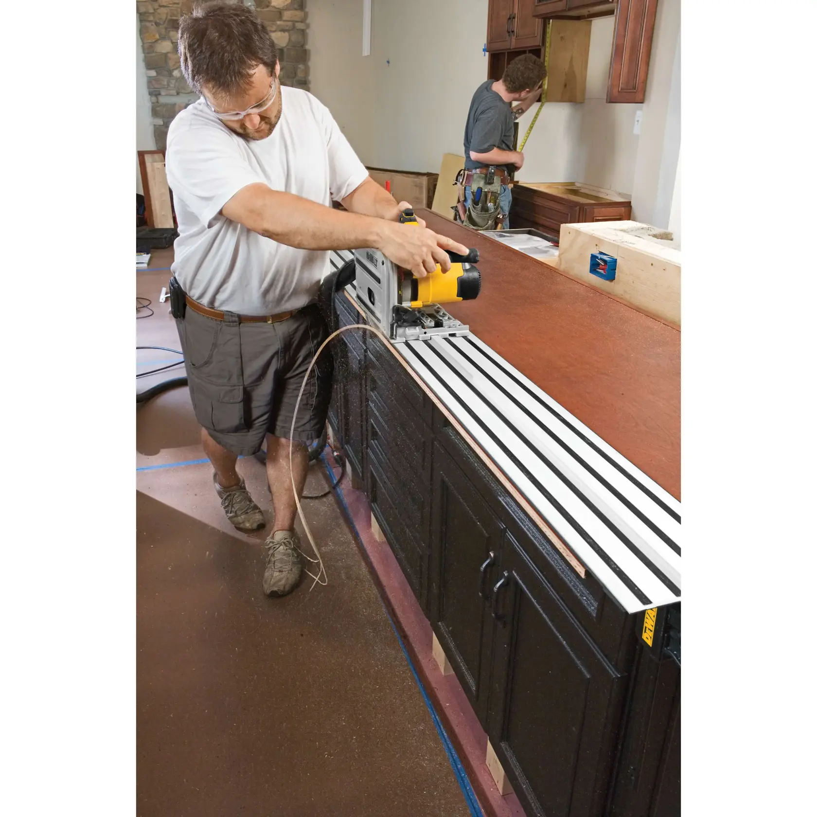 A person is using a DEWALT track saw (DWS520K) to cut a large wooden countertop in a kitchen setting. Another person in the background is measuring an area near the cabinets. The DEWALT saw is positioned on a guide rail for accurate cutting.
