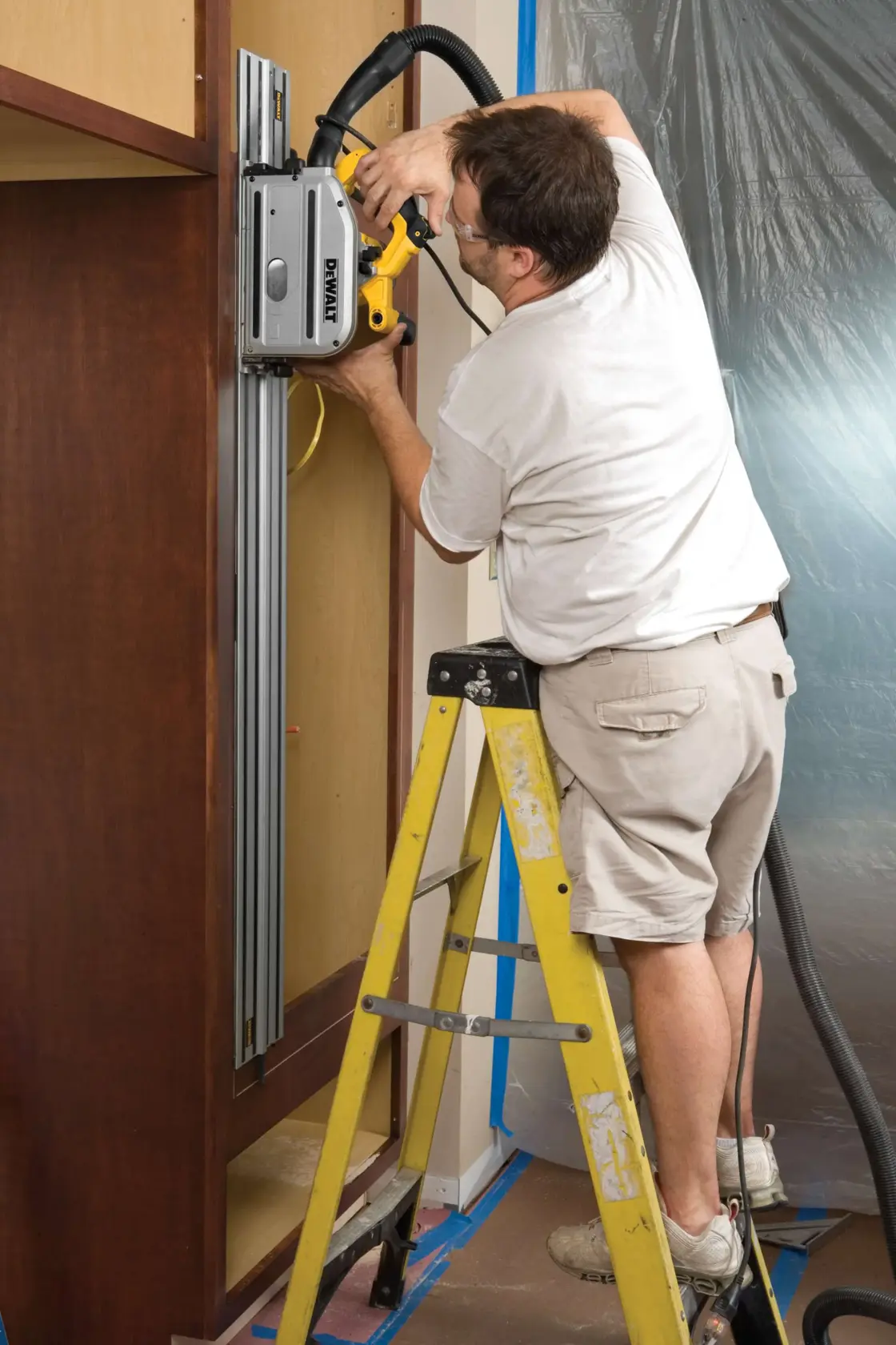 Person using a DEWALT DWS520K track saw on vertical wood panel while standing on a yellow step ladder indoors.