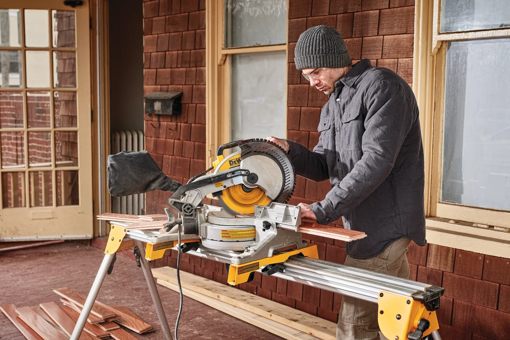 A person using a DEWALT DWS713 miter saw to cut wooden boards on a stand, wearing a jacket and beanie, in front of a building with brown shingles and windows.