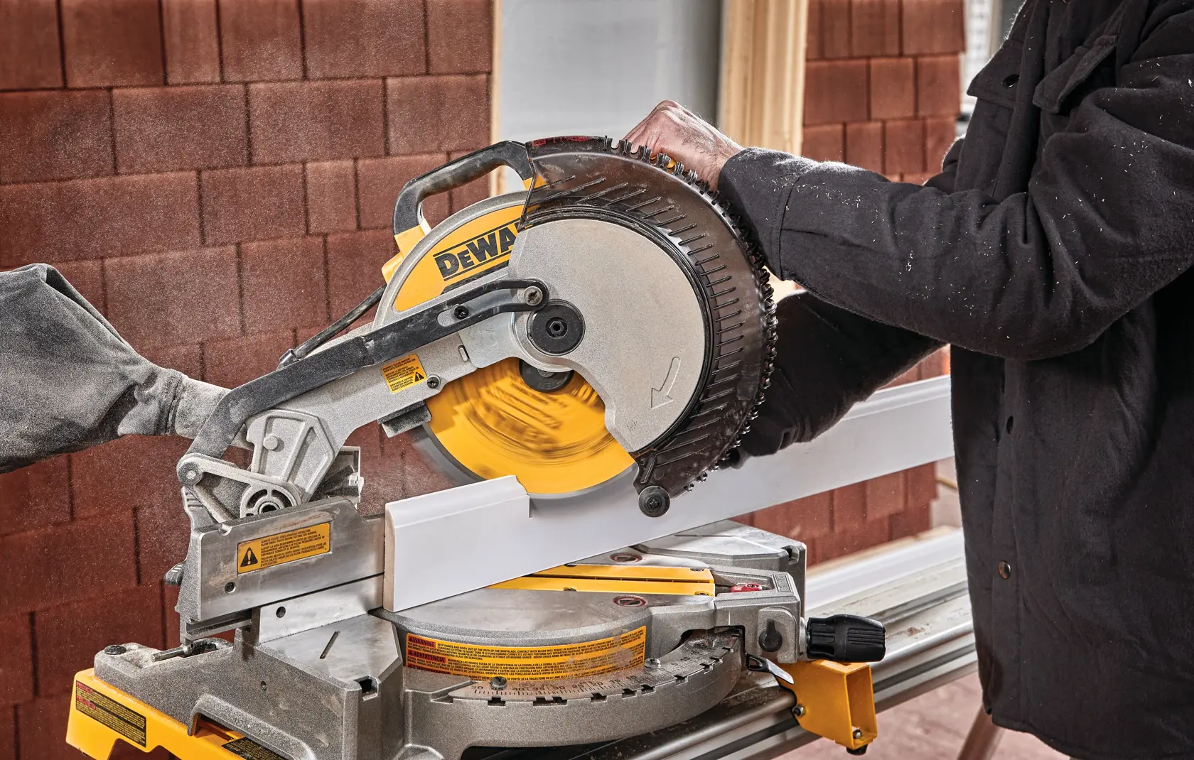 A person using a DEWALT DWS713 miter saw to cut a piece of white trim or molding. The saw is mounted on a workbench and dust is visible in the air.
