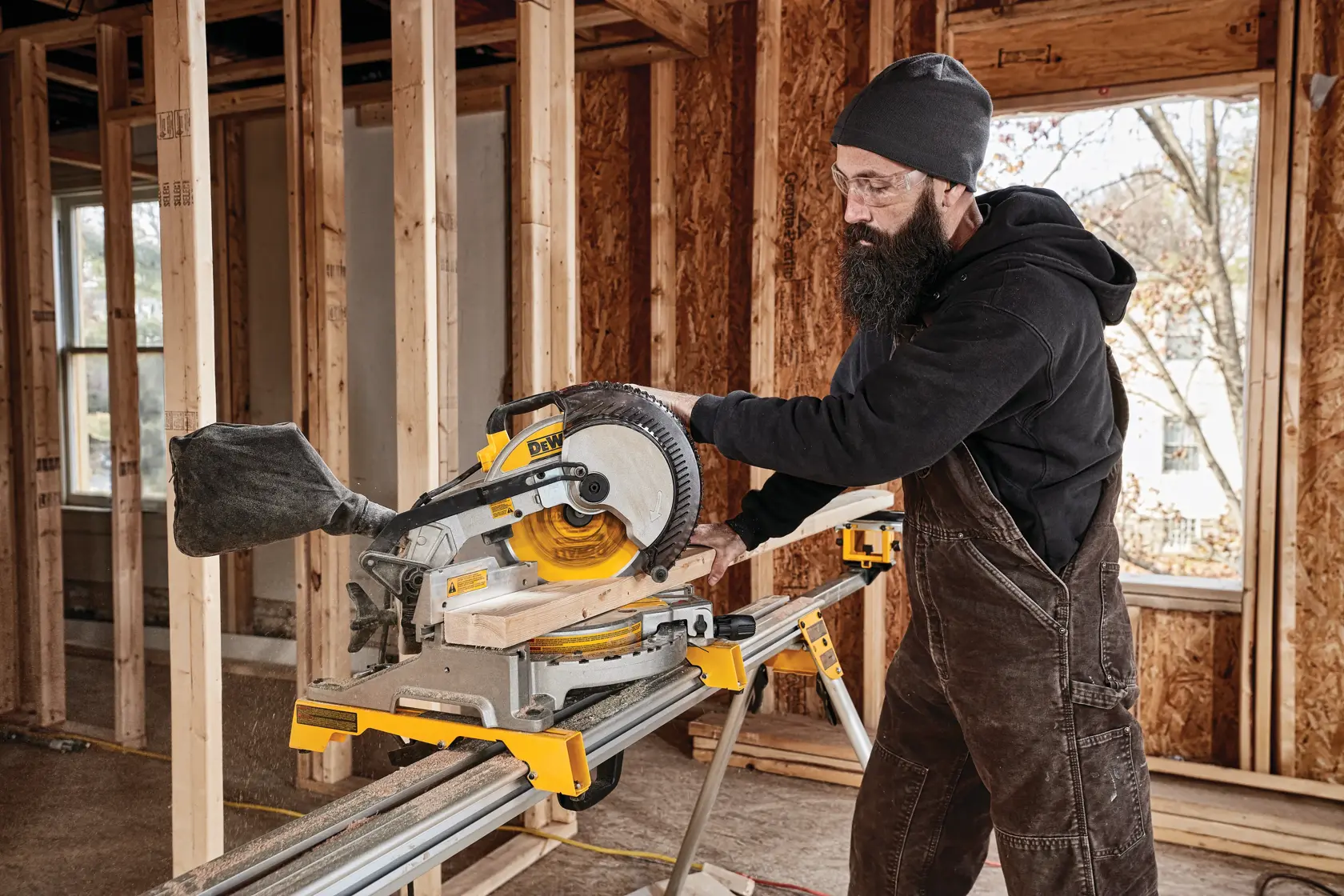 A person wearing work overalls and a hat is operating a DEWALT DWS713 miter saw to cut a piece of wood inside a partially constructed building.