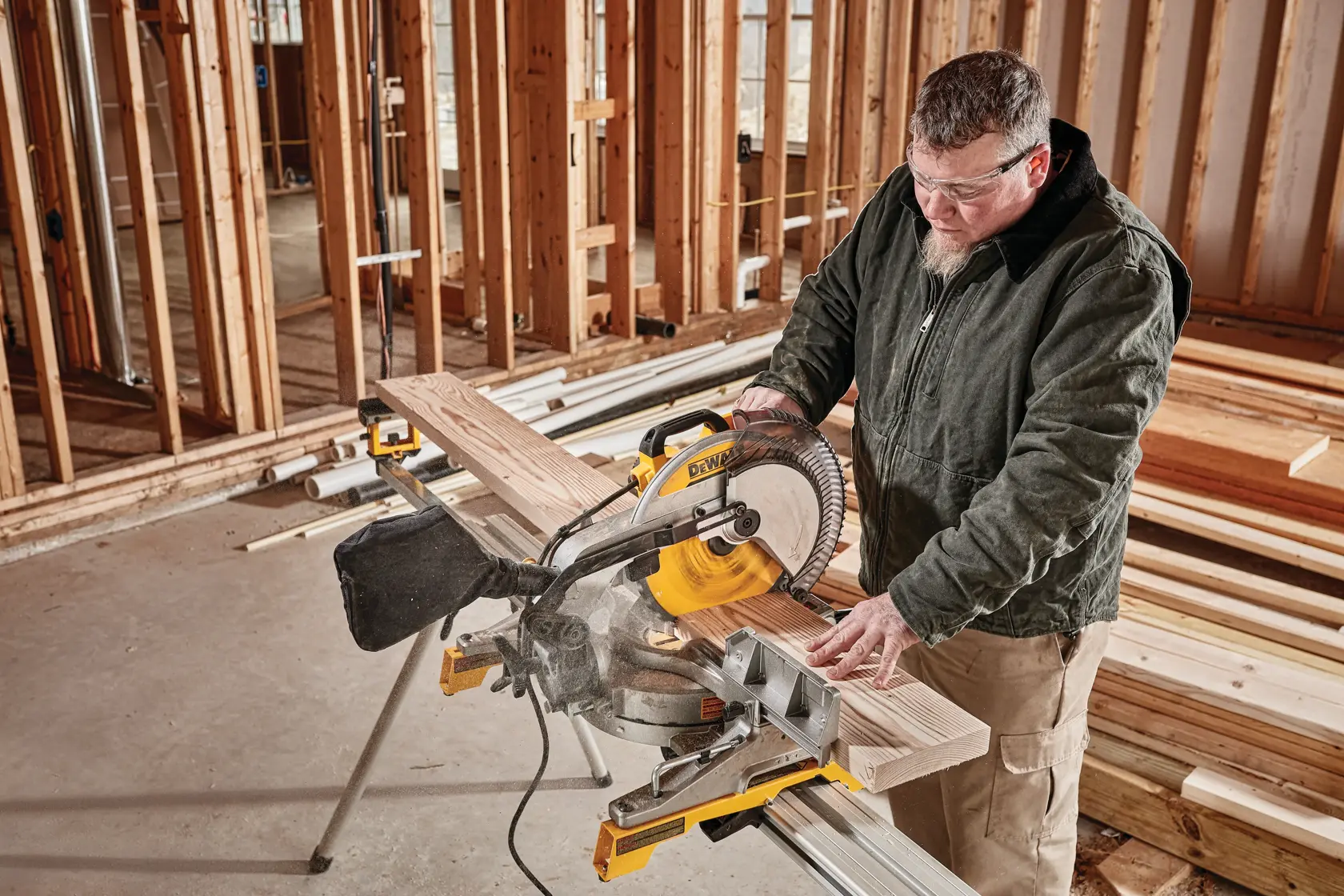 A person uses a DEWALT DWS715 miter saw to cut a wooden plank at a construction site with unfinished wooden framing and stacks of lumber in the background.