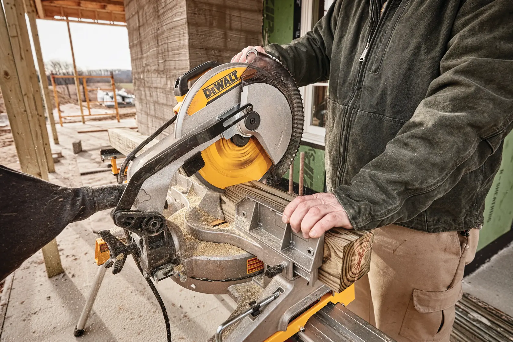 A person using a DEWALT miter saw to cut a piece of wood outdoors, with construction materials and framework visible in the background.