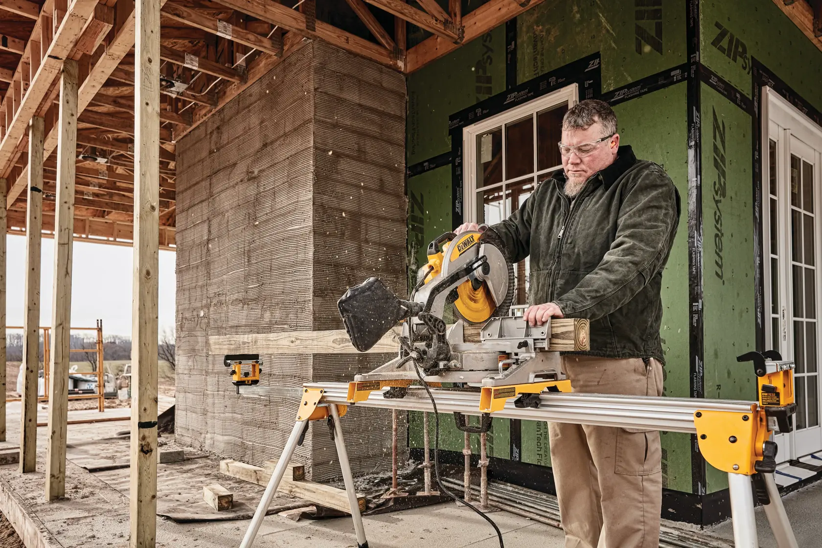 A person using a DEWALT DWS715 miter saw to cut a piece of wood on a construction site. The saw is mounted on a stand, and there is an unfinished building structure in the background.
