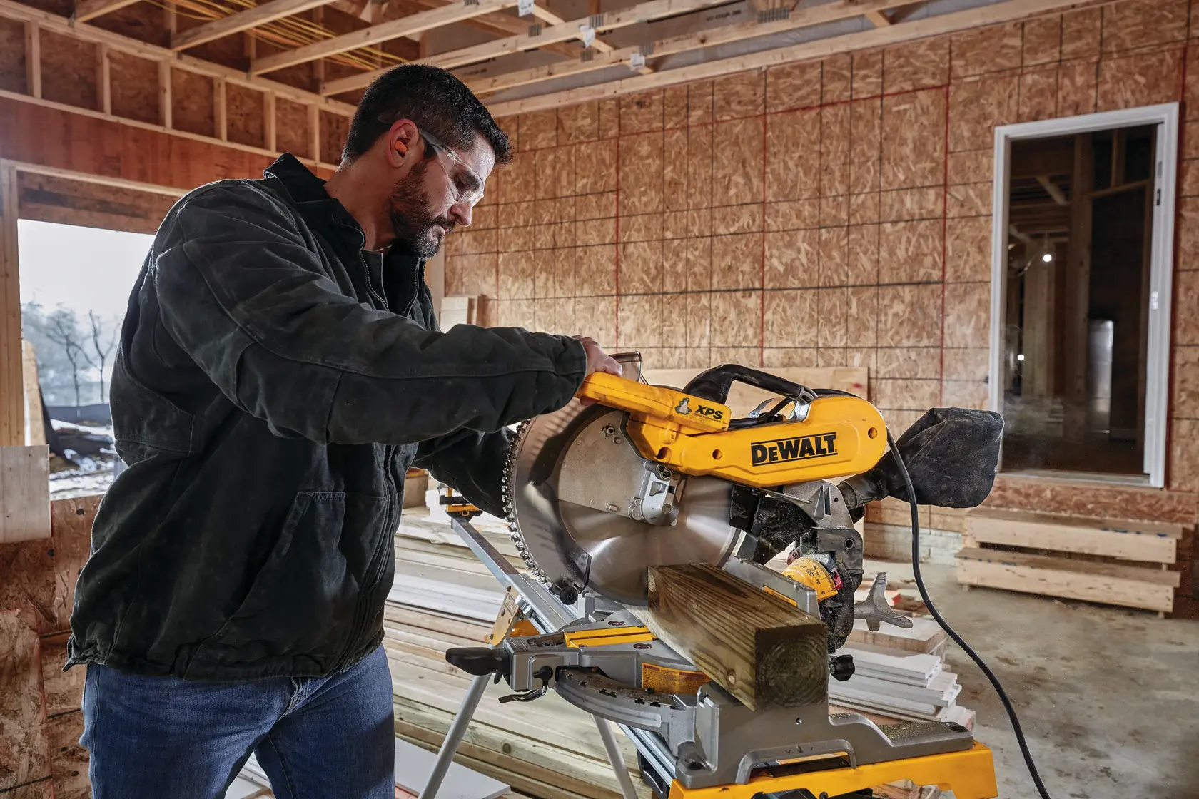 A person using a DEWALT DWS716XPS miter saw to cut a piece of wood inside a construction site with wooden framing and unfinished walls.