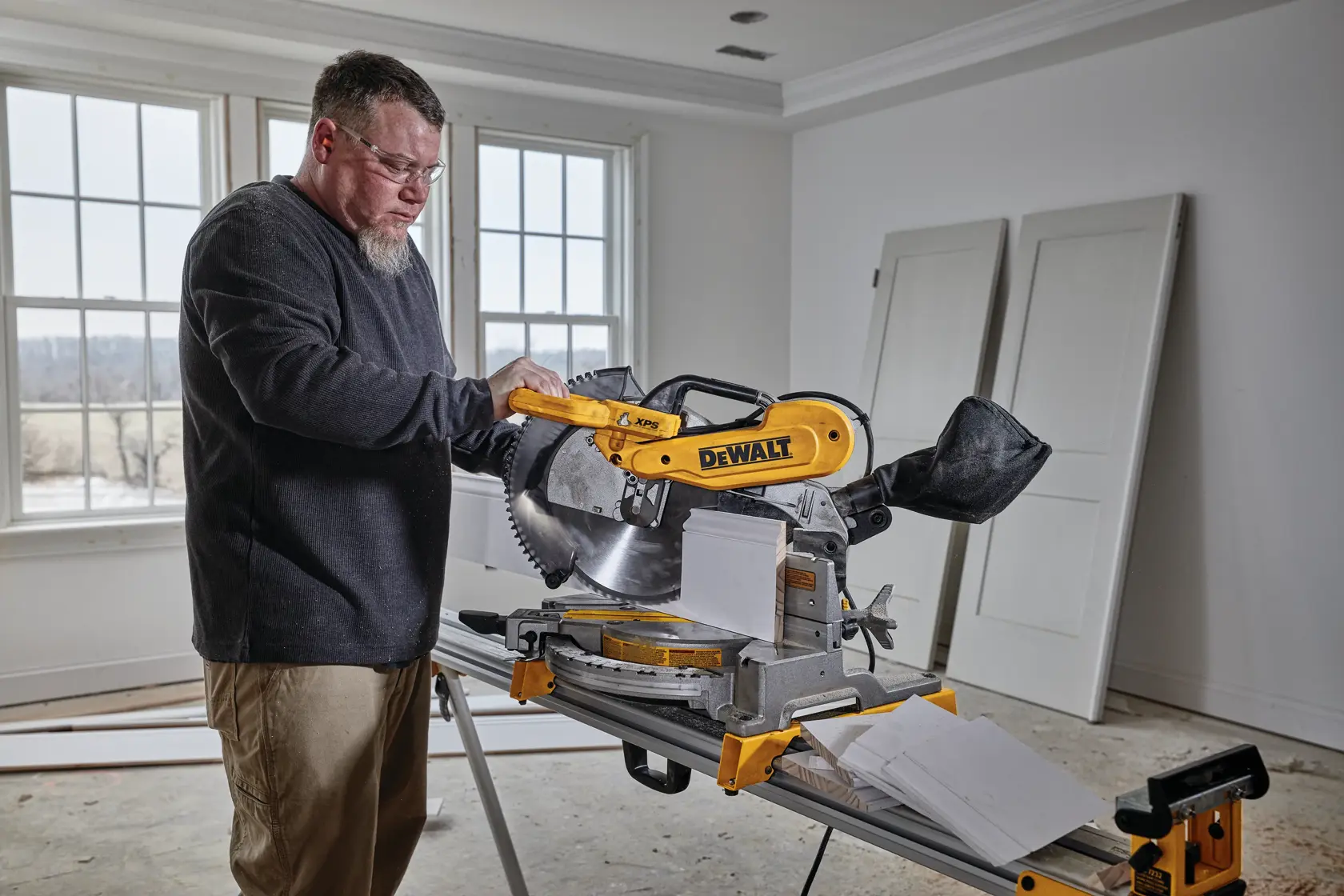 A person operating a DEWALT miter saw (model DWS716XPS) inside a room with construction materials and doors in the background.