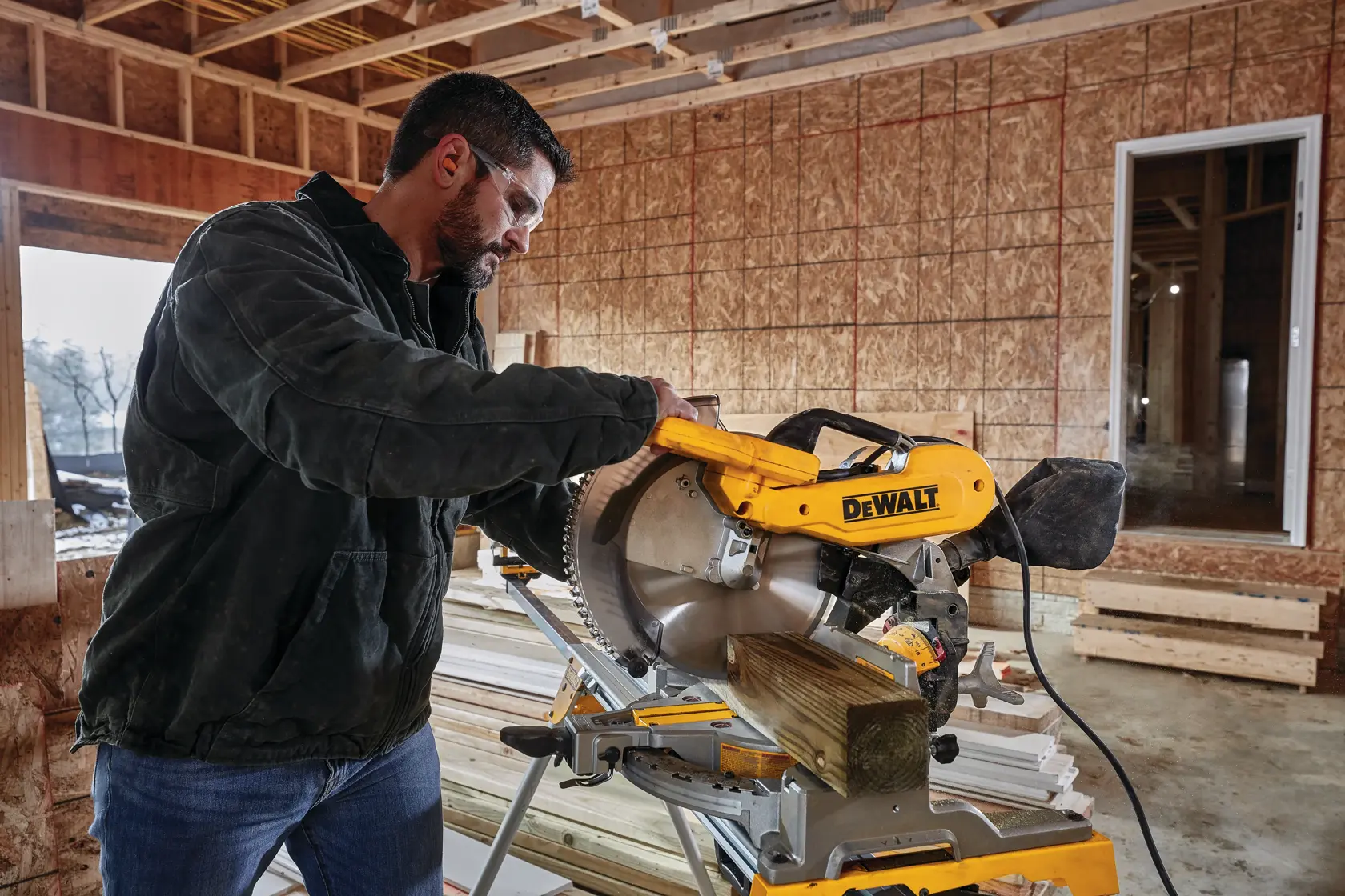 A person using a DEWALT miter saw to cut a wooden plank inside a construction site with exposed wall framing.