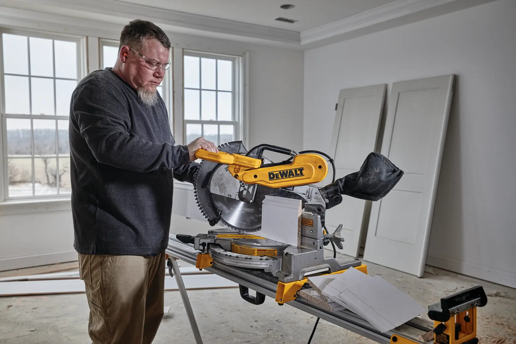 A person uses a DEWALT DWS716 miter saw to cut a piece of wood in a room with large windows and two white doors leaning against the wall. The individual's face is blurred for privacy.