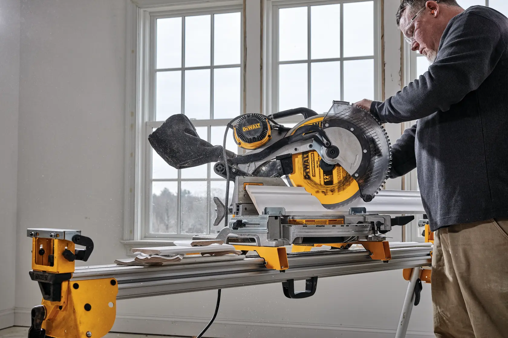 A person uses a DEWALT DWS716 miter saw to cut wooden boards, with the saw placed on a yellow stand in a bright room with large windows.