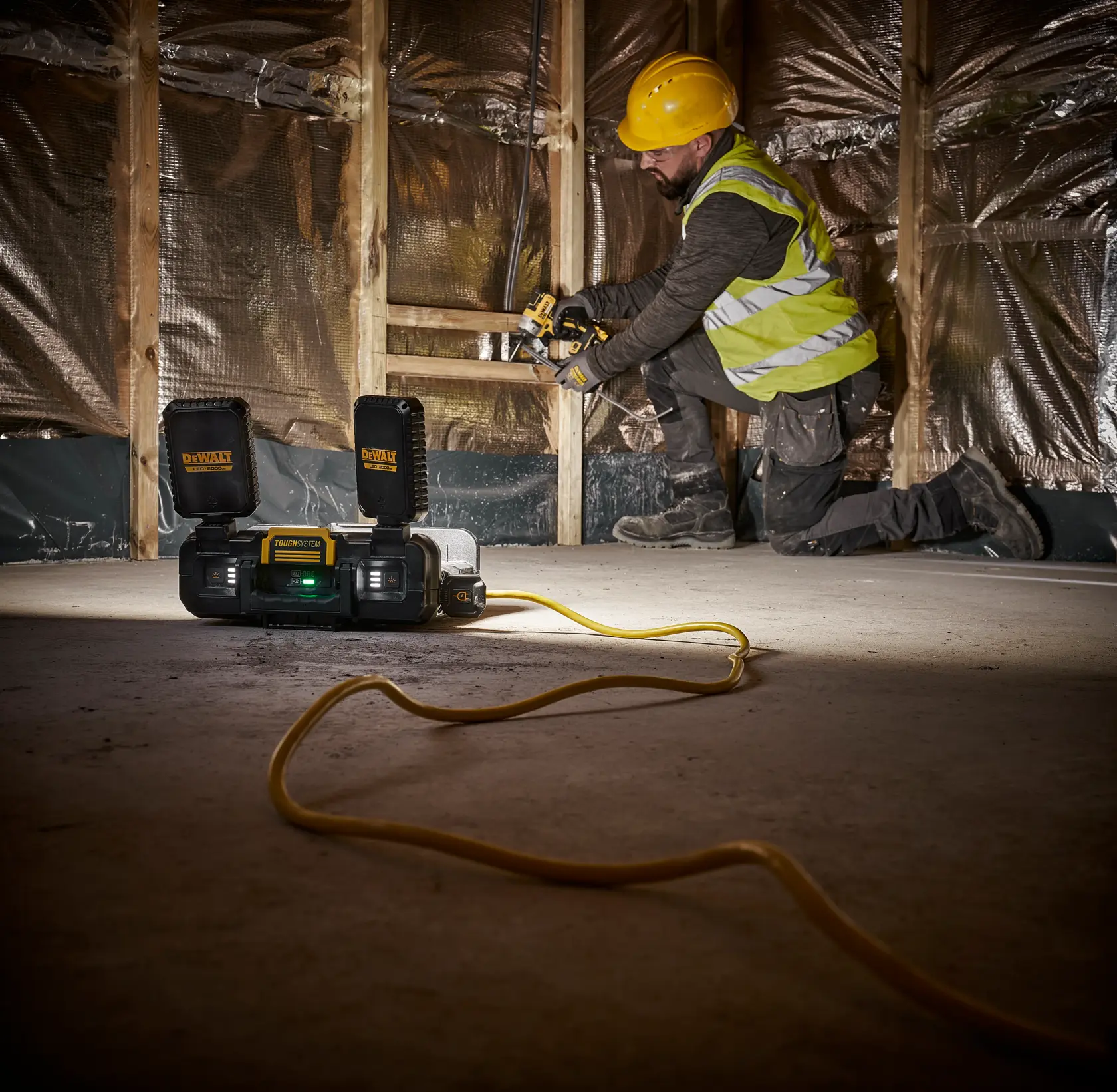 A DEWALT Power Station DWST08060 with two battery packs is placed on the floor of a construction site. A person wearing a yellow hard hat and safety vest is working on wooden framing in the background. An extension cord is visible stretching across the floor.