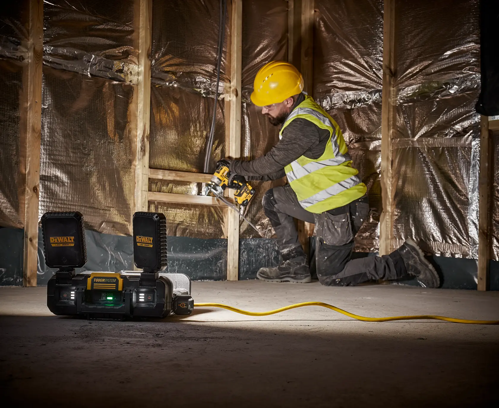 A construction worker wearing a yellow hard hat and reflective vest is kneeling while using a DEWALT power tool in a partially finished room with wooden framing and insulation. In the foreground, a DEWALT portable work light and power station (model DWST08060) with batteries and a cord is placed on the floor, illuminating the workspace.