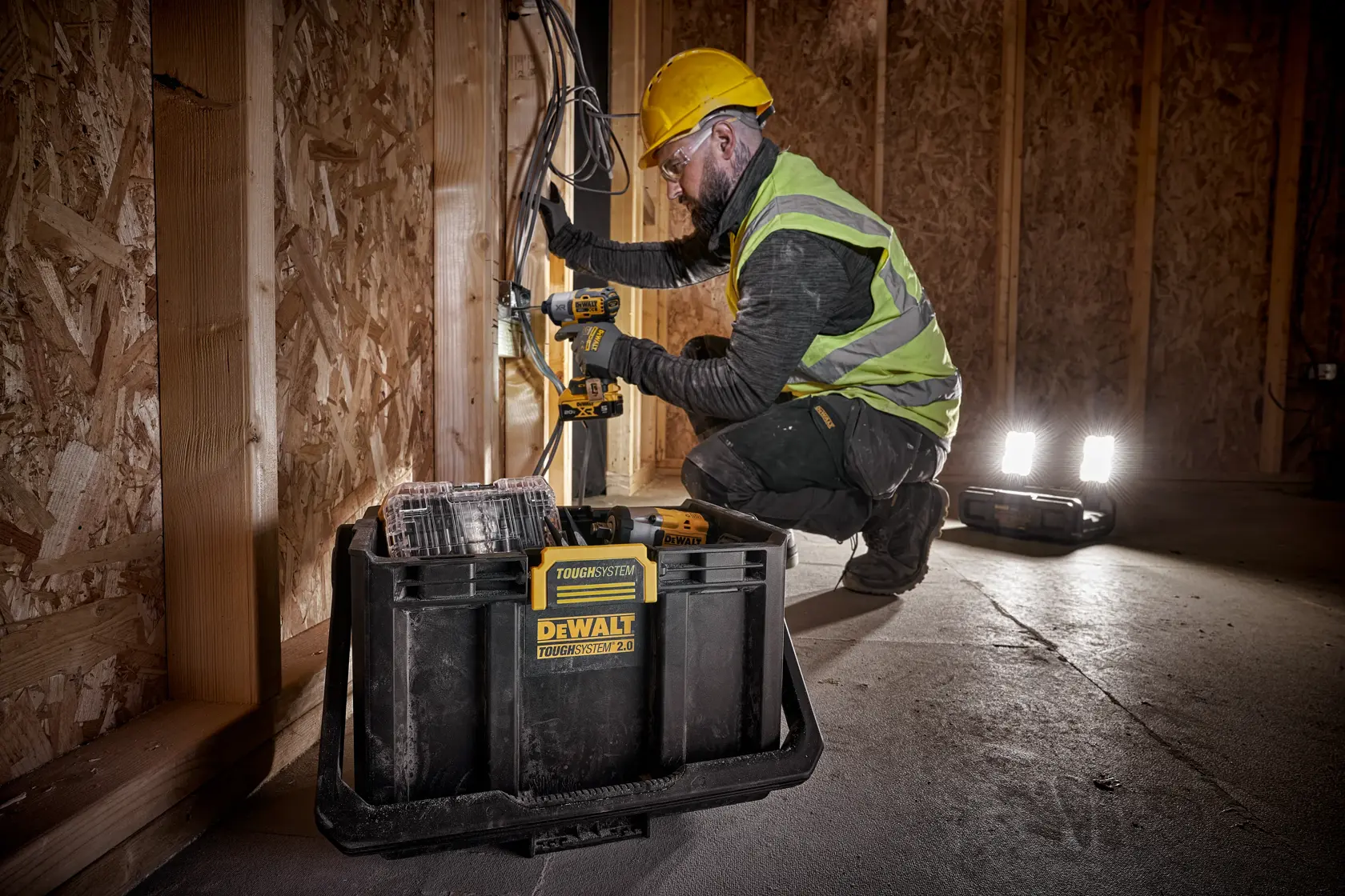 A construction worker wearing a yellow hard hat and a reflective vest is kneeling inside a wooden framed building, using a cordless power drill. In the foreground, a DEWALT ToughSystem 2.0 toolbox (model DWST08060) is visible on the floor. The workspace is illuminated by a portable work light.