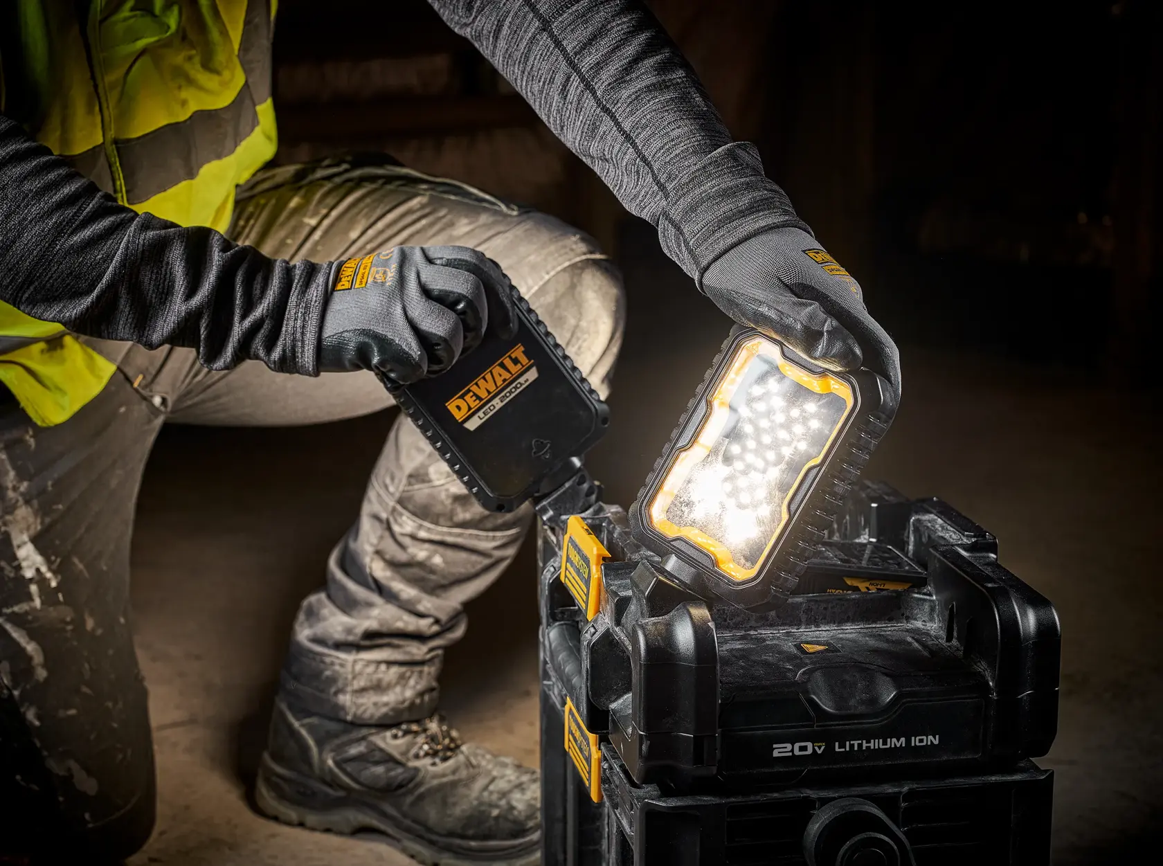 A person wearing work gloves and a high-visibility vest is adjusting a DEWALT LED work light attached to a black stackable toolbox system. The toolbox has a 20V lithium-ion battery and is designed for use on construction sites.