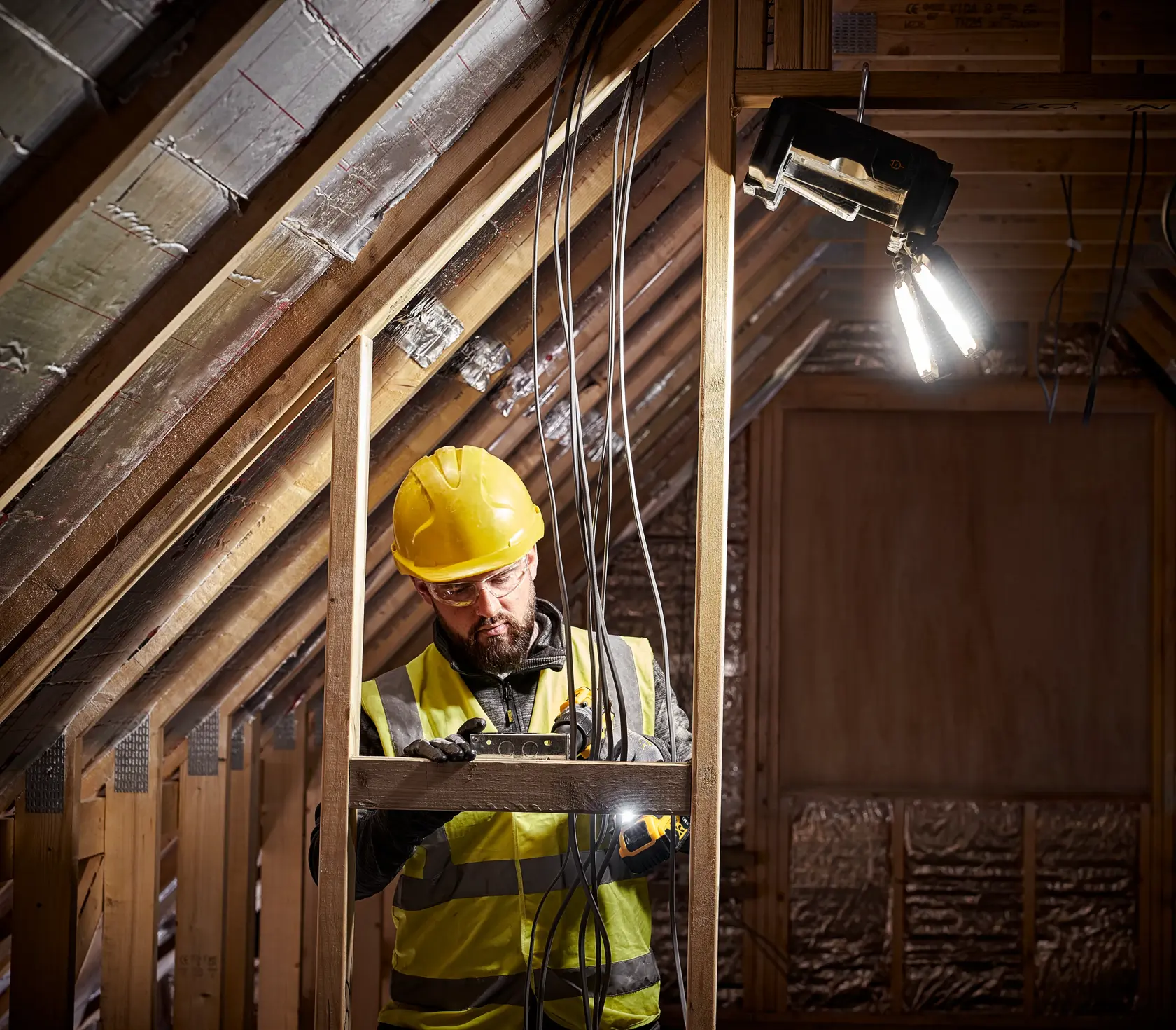 A construction worker wearing a yellow hard hat and reflective vest is using a DEWALT tool to work on electrical wiring inside an attic. There is a bright work light attached to the wooden beams above.