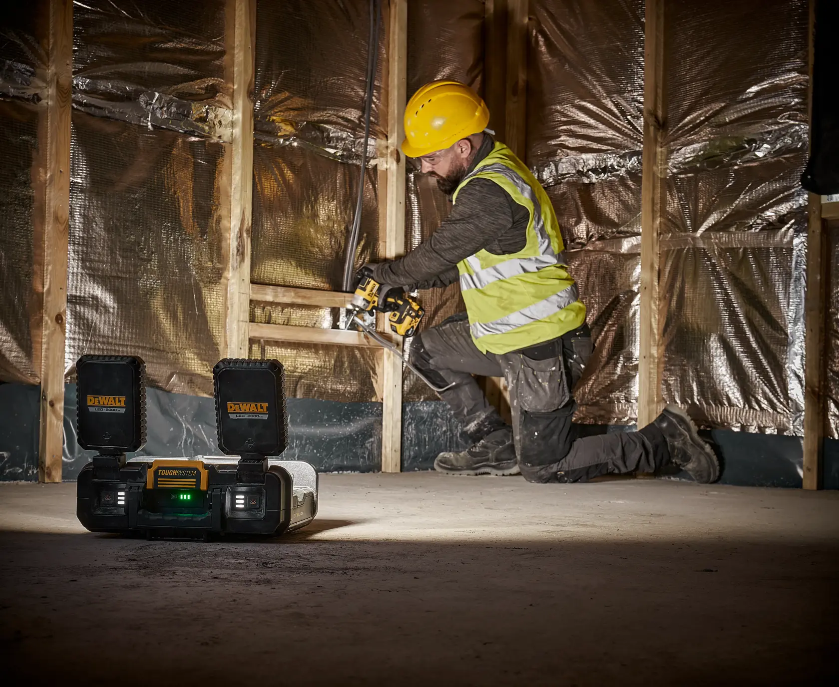 A construction worker wearing a yellow hard hat and reflective vest is kneeling and operating a power tool near a wooden frame wall with insulation. In the foreground, a DEWALT ToughSystem radio is placed on the floor with its compartments open. The scene takes place indoors on a construction site.