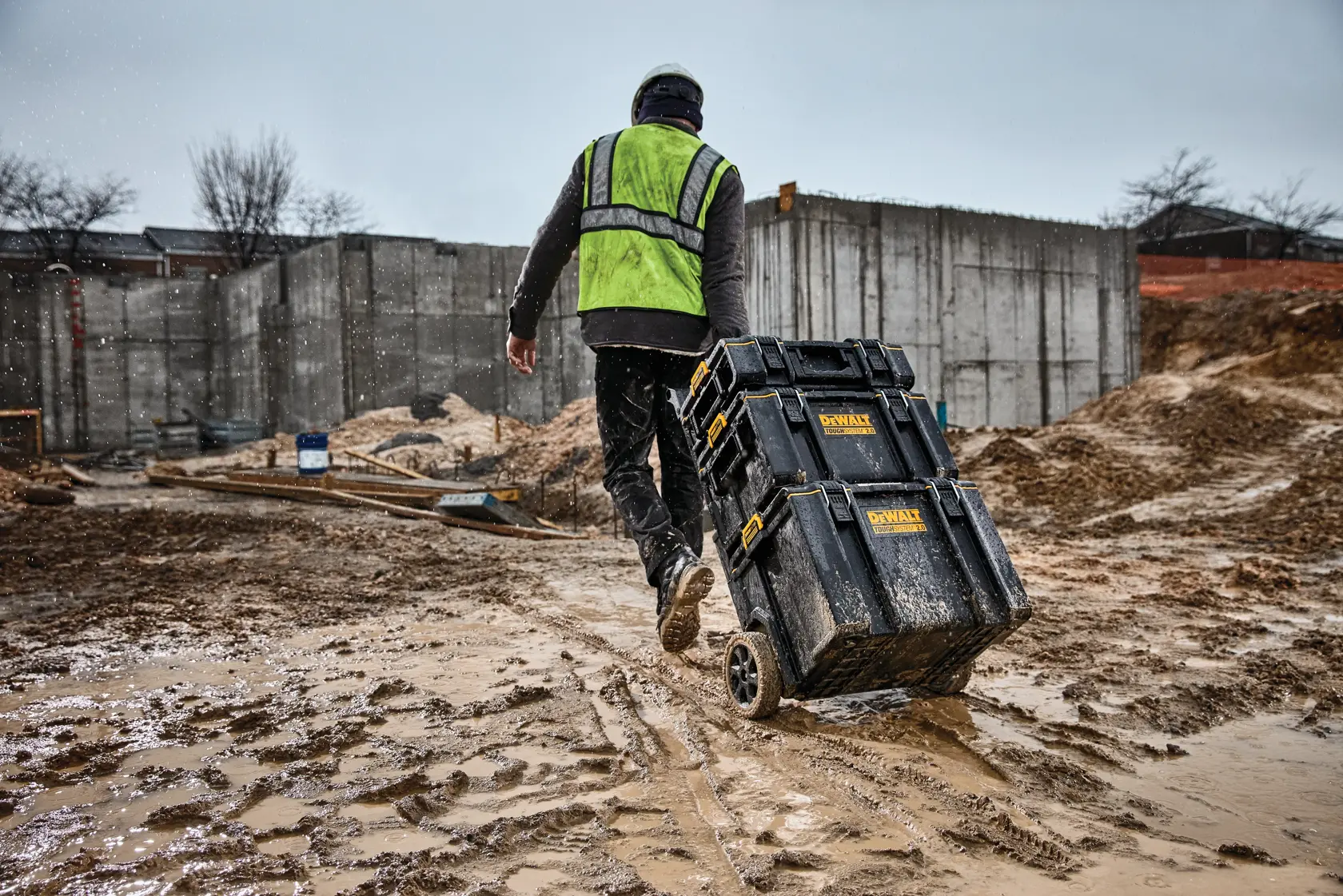 A person wearing a green safety vest and helmet pulls a stack of DEWALT tool boxes on wheels across a muddy construction site.