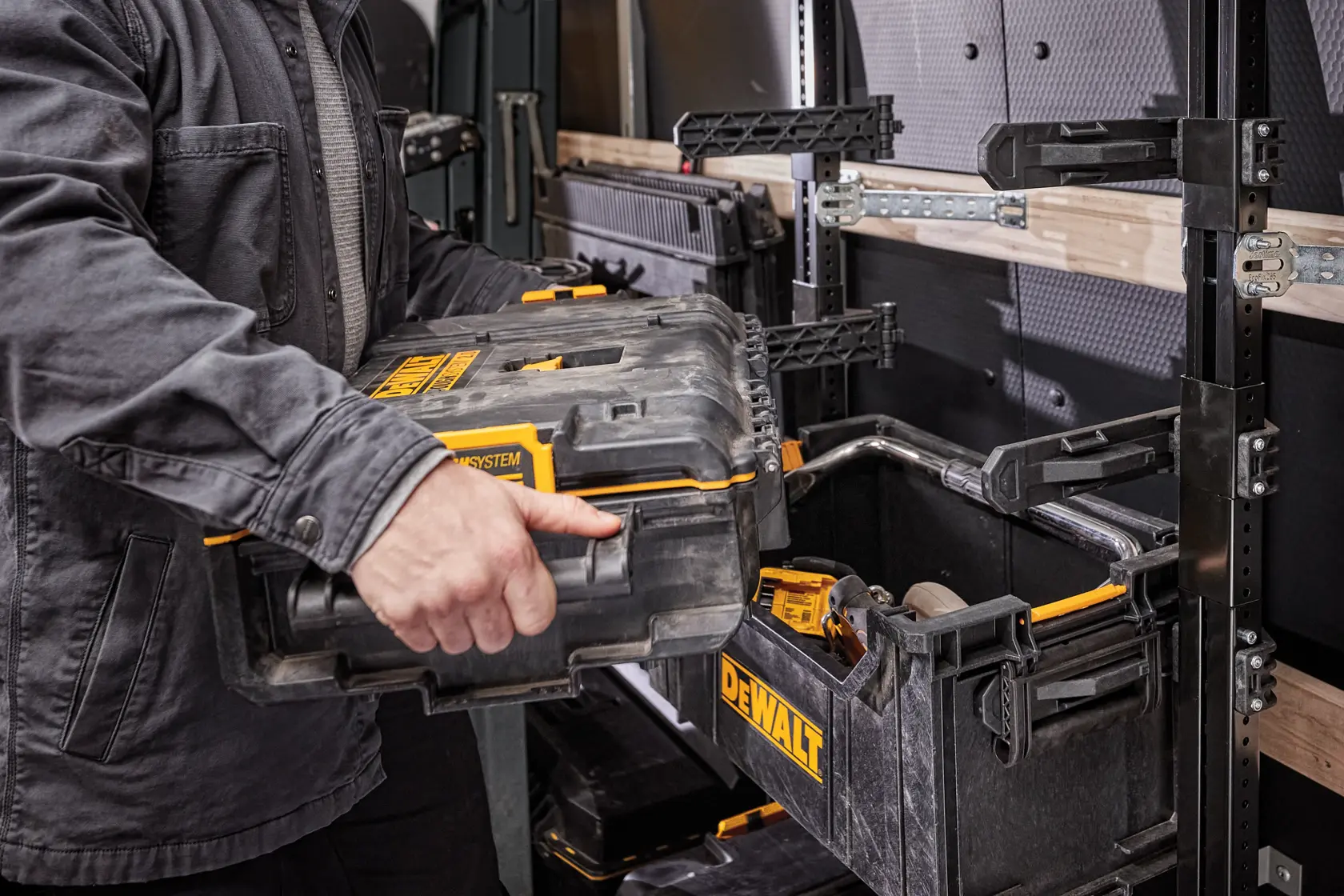 A person is placing a DEWALT ToughSystem toolbox into a storage rack in a workshop or vehicle. The toolbox and rack are black with yellow accents and are designed for organizing and transporting tools.