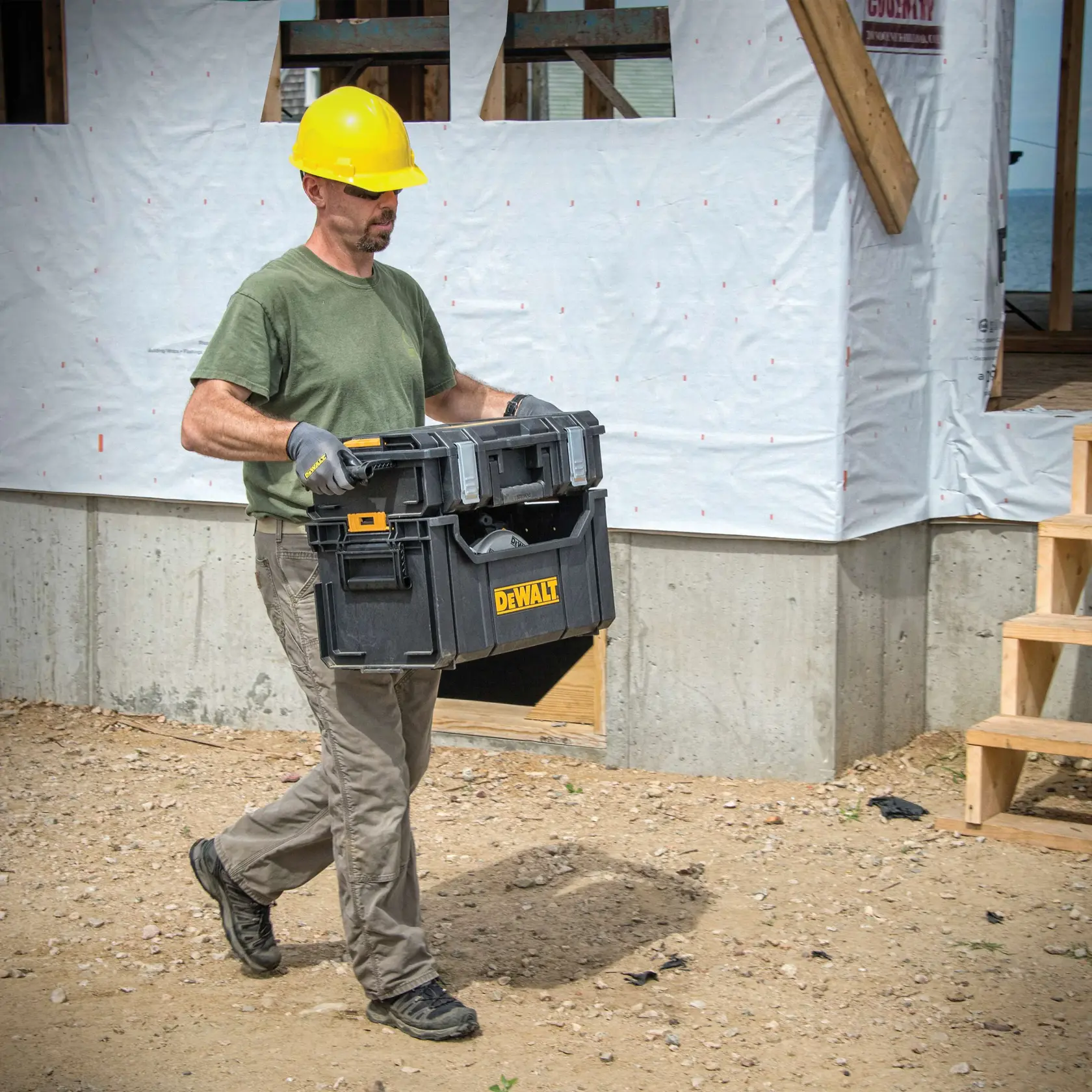 A person wearing a yellow hard hat, gloves, and a green t-shirt is carrying a black DEWALT tool box (SKU: DWST08205) at a construction site with a partially built structure in the background.
