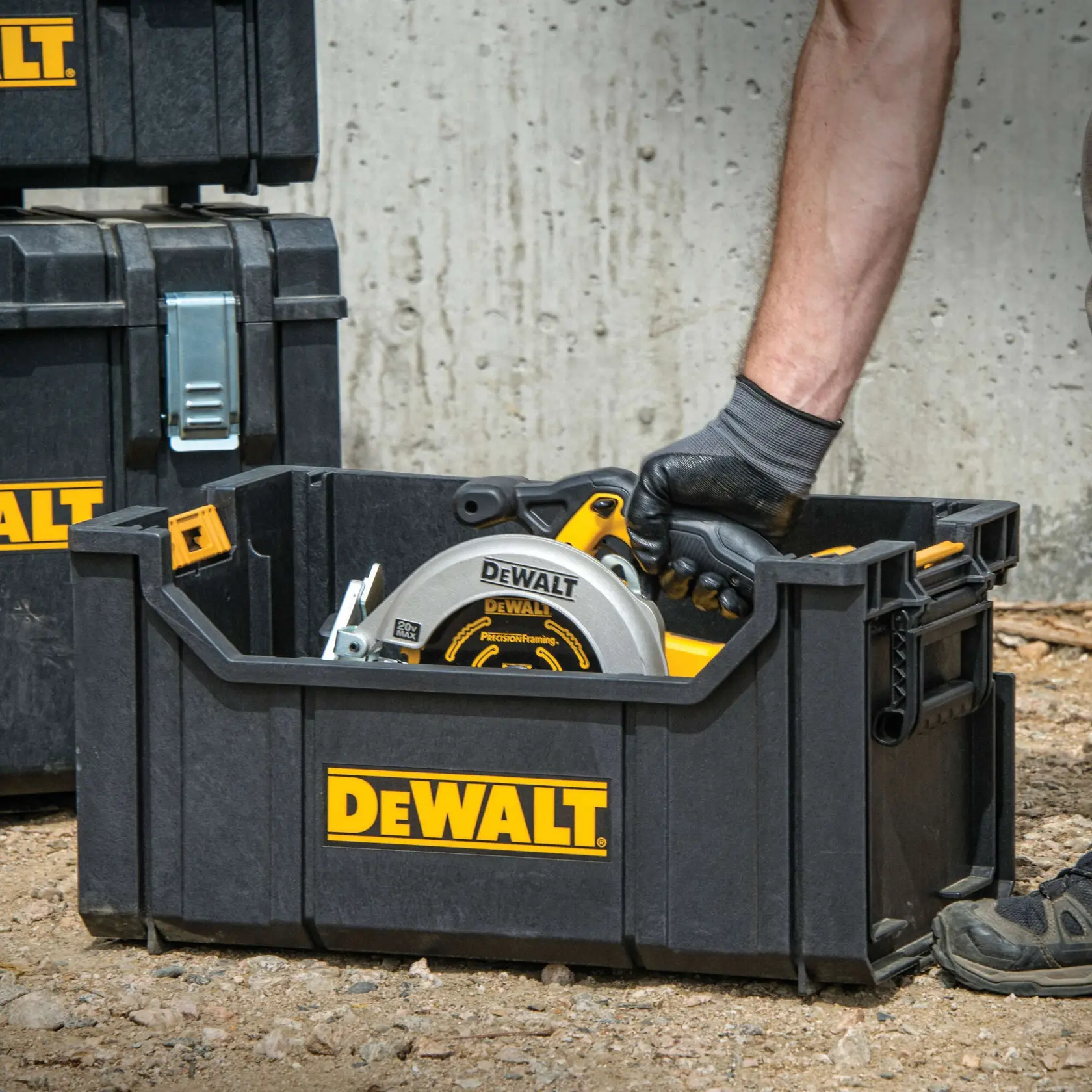 A DEWALT branded black storage box is shown on a construction site. A person wearing gloves is placing a circular saw and other tools inside the box. Additional DEWALT storage containers are visible in the background.