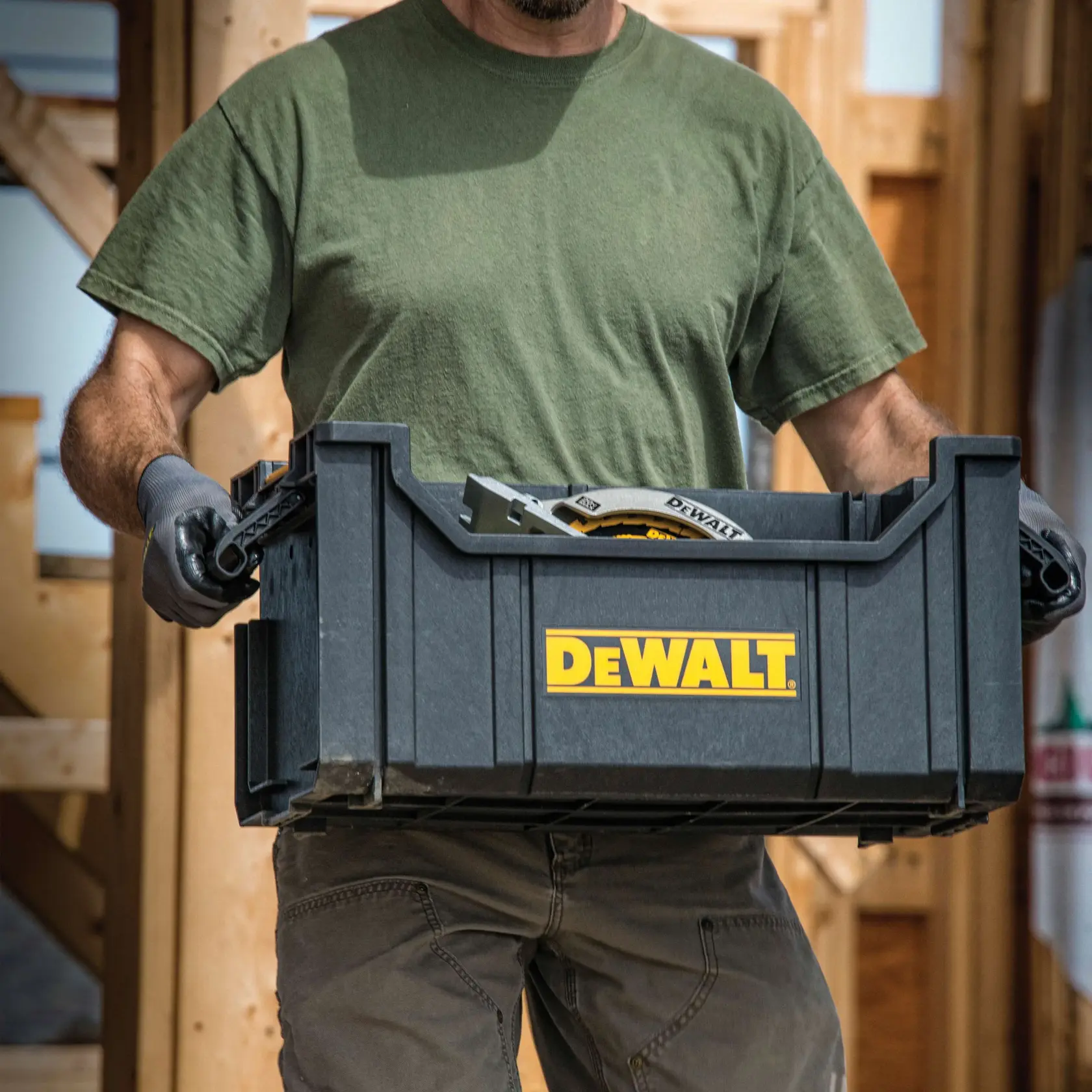A person wearing a green shirt and gloves is carrying a black DEWALT plastic storage container filled with tools. The DEWALT logo is prominently visible on the front of the container. The scene appears to be at a construction site with wooden framing in the background.