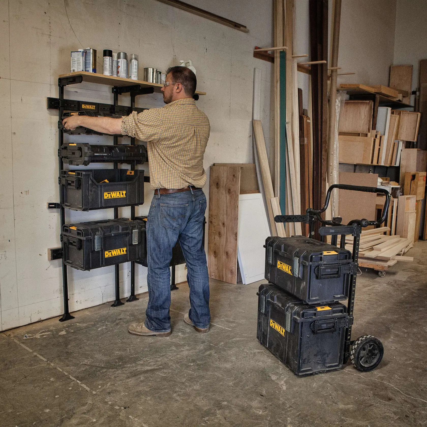 A person is organizing DEWALT storage boxes on a wall-mounted rack in a workshop. Additional DEWALT storage boxes are stacked on a wheeled cart nearby. The workshop has various wooden boards and shelves in the background.