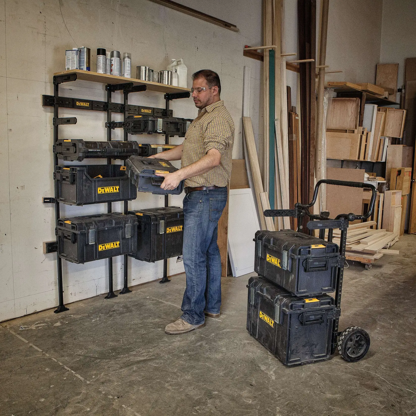 A man in a workshop is organizing DEWALT stackable toolboxes, placing one onto a wall-mounted storage rack. Several black and yellow DEWALT toolboxes are stored on the rack, and two are stacked on a wheeled cart nearby. The workspace contains wooden boards and shelves with various supplies.