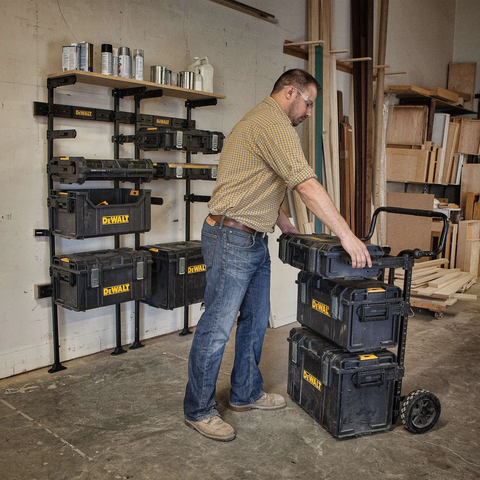 A person in a workshop organizes DEWALT storage boxes, some mounted on a wall rack and others stacked on a rolling cart. The workshop contains wood materials and shelves in the background.