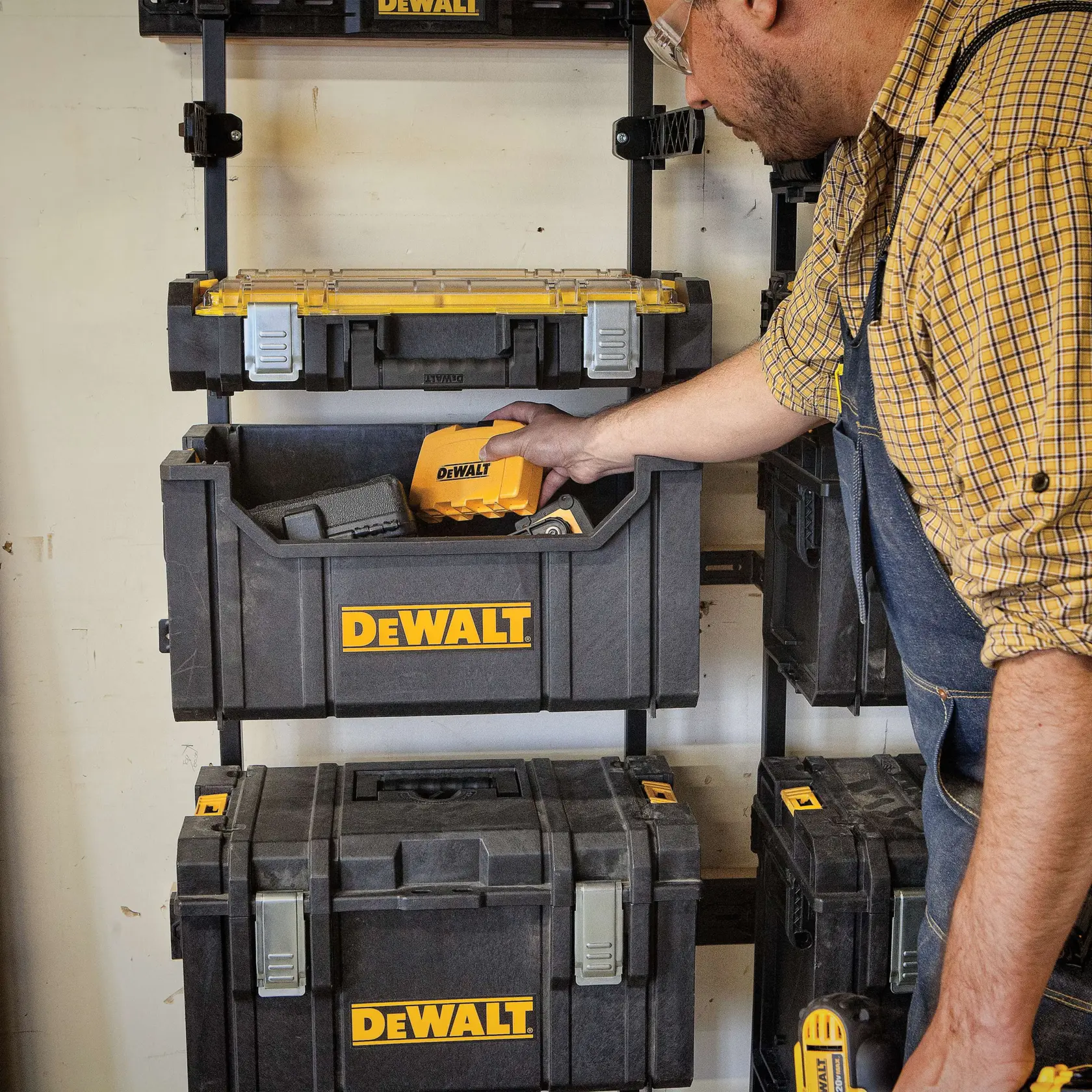 A person in a plaid shirt and overalls is organizing DEWALT branded storage boxes mounted on a wall. The person is placing a DEWALT power tool inside one of the boxes. The storage boxes are black with yellow DEWALT logos.