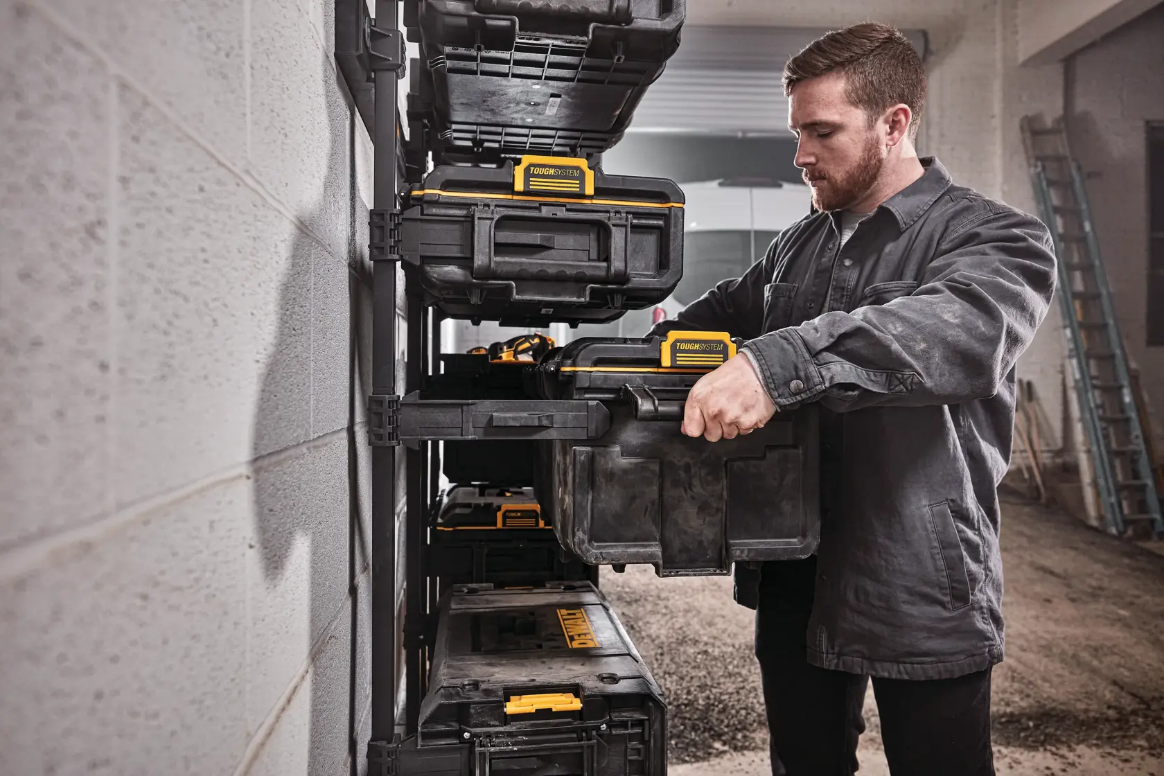 A person is organizing DEWALT storage boxes on a wall-mounted rack in a workshop or garage. The individual's face is blurred and they are wearing a dark work jacket.