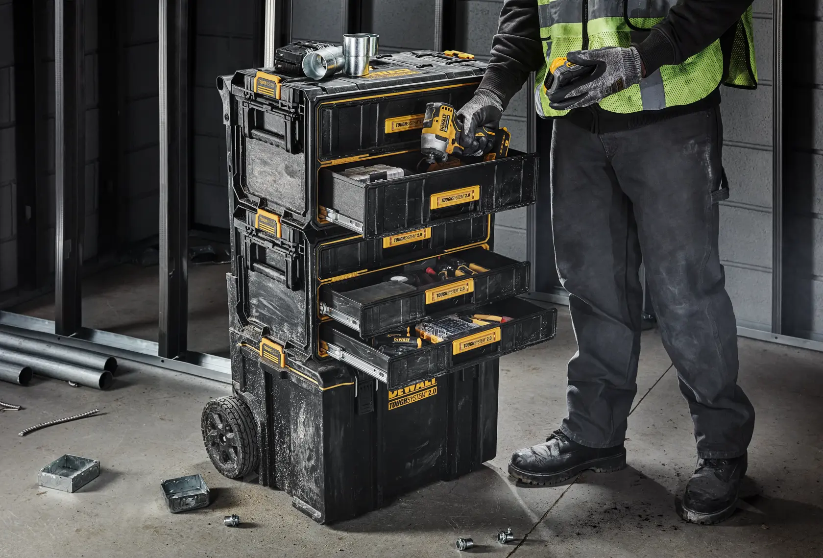 A worker wearing gloves and a safety vest is standing next to a DEWALT rolling tool storage unit with several open drawers filled with tools. The unit is on wheels and appears to be used in a construction setting.