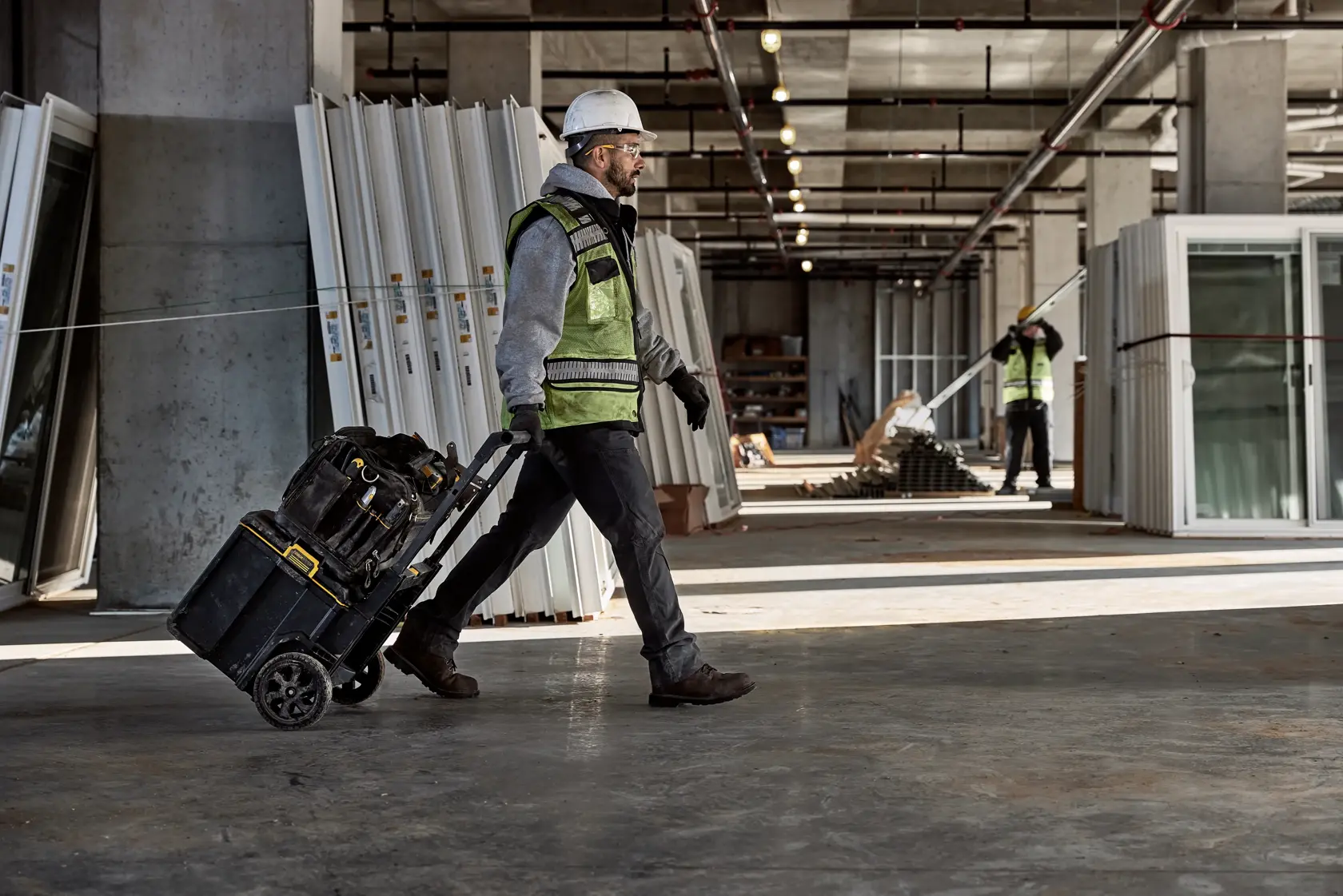 A construction worker wearing safety gear pulls a rolling DEWALT tool box (SKU: DWST08350) across a building site with large panels and windows stacked along the walls.