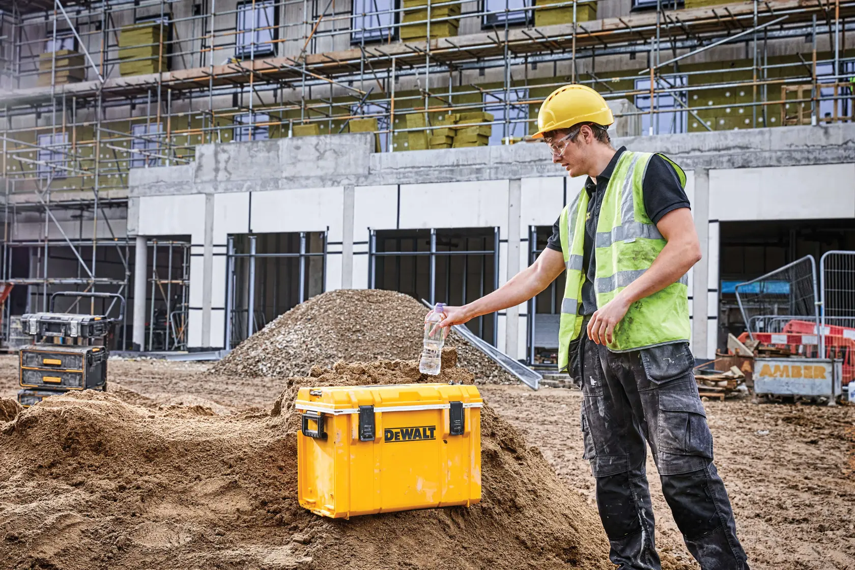 A yellow DEWALT storage box (SKU: DWST08404) placed on a pile of dirt at a construction site, with a worker wearing a safety vest and a yellow hard hat standing next to it holding a plastic water bottle. Scaffolding and building materials are visible in the background.