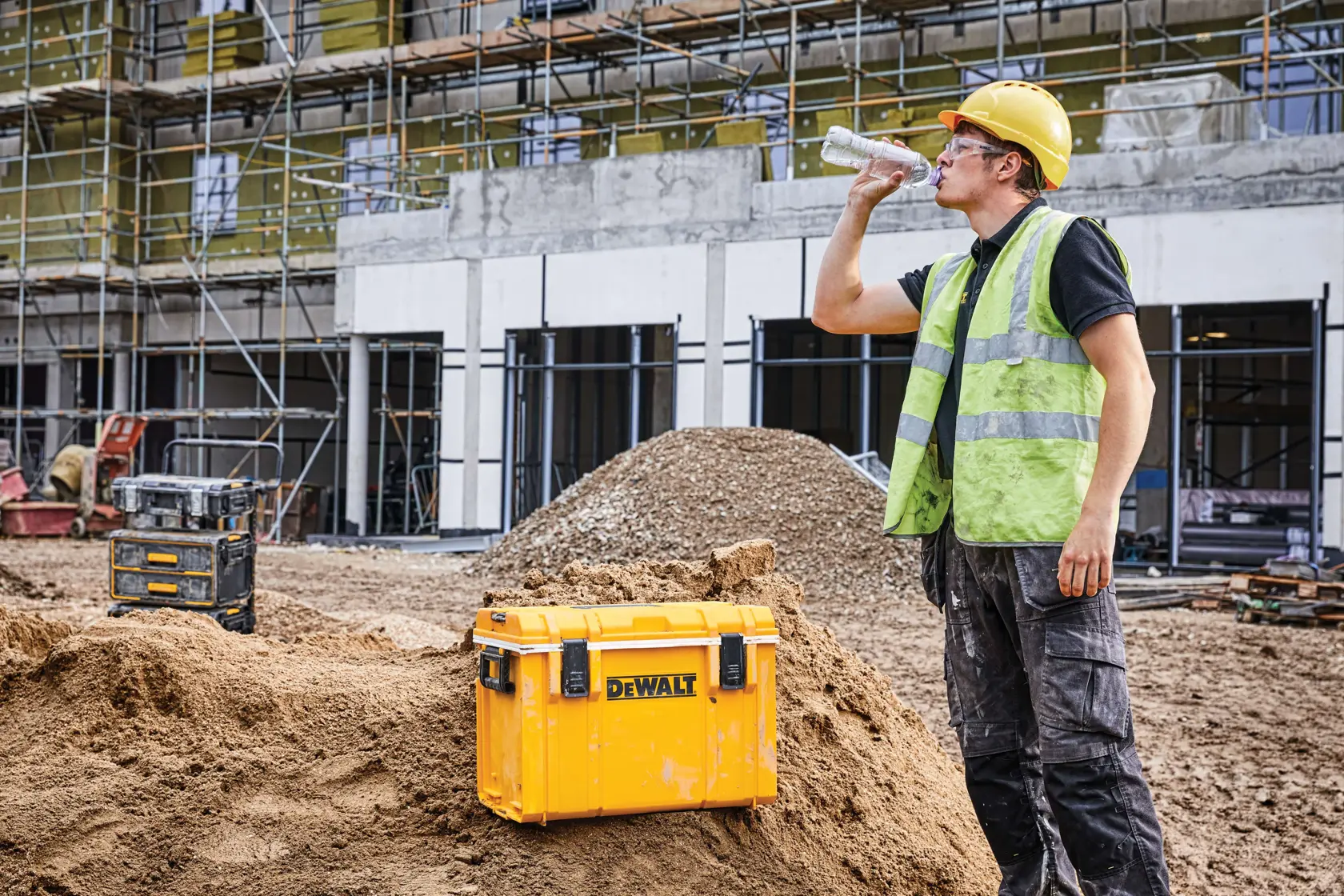 A DEWALT DWST08404 yellow toolbox is placed on a dirt mound at a construction site. A person wearing a hard hat and safety vest is drinking from a bottle, with building scaffolding and another toolbox visible in the background.