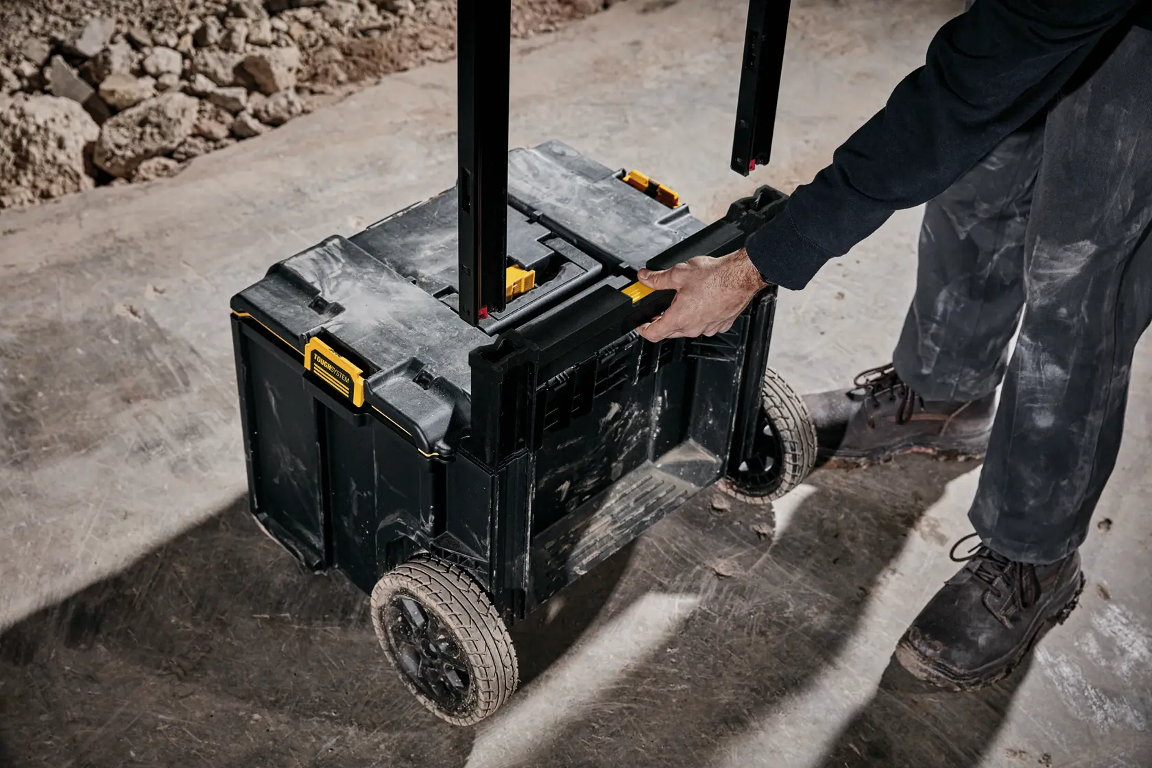 A person handling a DEWALT rolling tool storage box with large wheels and a telescopic handle on a construction site.
