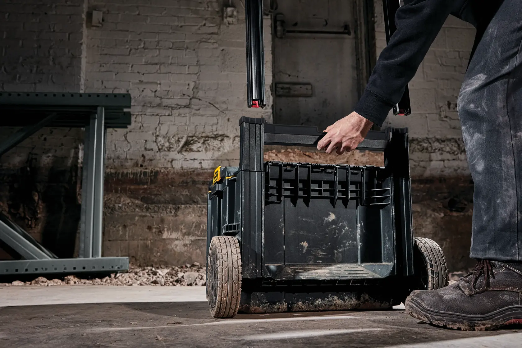 A person is pulling the handle of a DEWALT black rolling toolbox with large wheels, positioned on a dirty workshop floor against a brick wall.