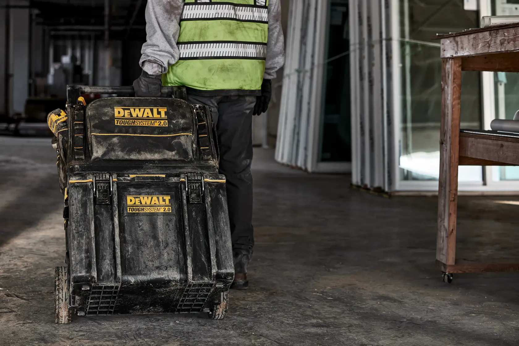A person wearing a safety vest and gloves is pulling a DEWALT ToughSystem 2.0 rolling tool box in an industrial indoor setting with construction materials and work tables around.