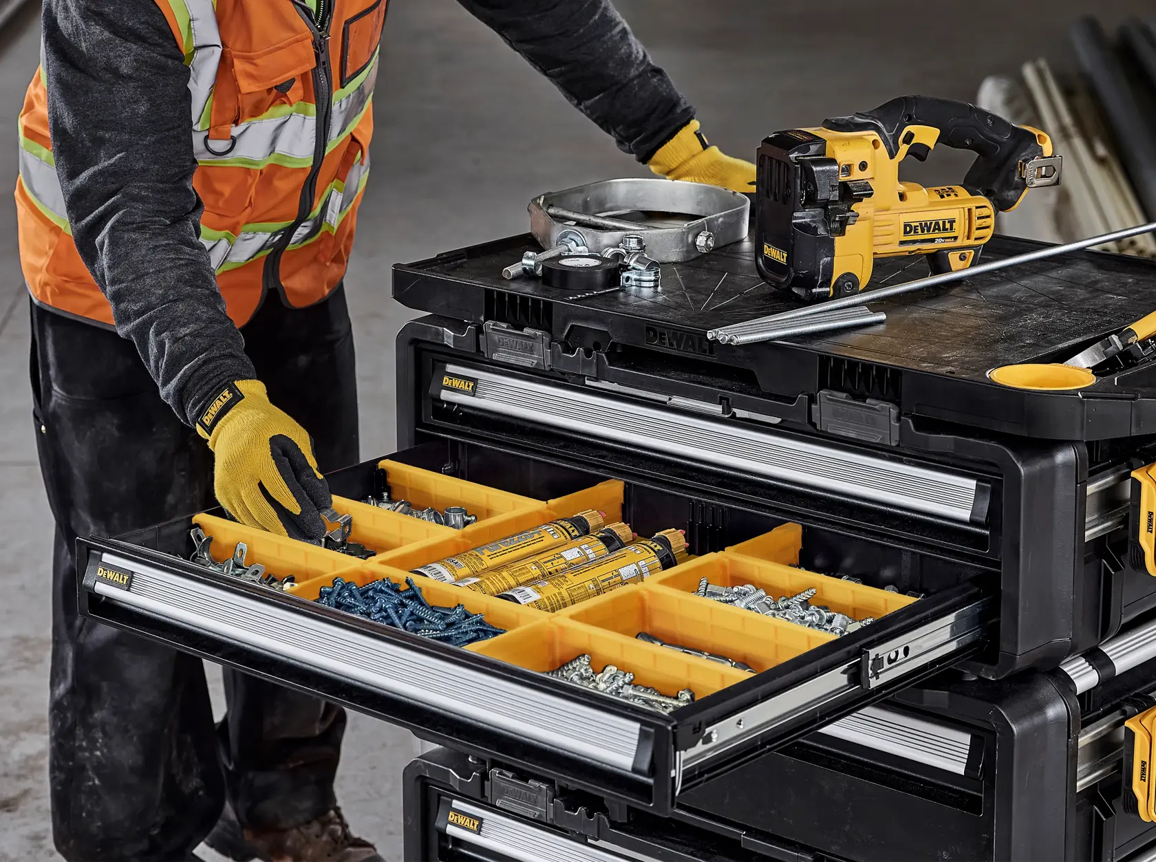 A person wearing work gloves and a high-visibility vest is organizing items in a DEWALT stackable storage drawer cabinet, which contains yellow compartment trays filled with screws, bolts, and other hardware. On top of the cabinet, there is a DEWALT power tool, metal parts, and a steel rod.