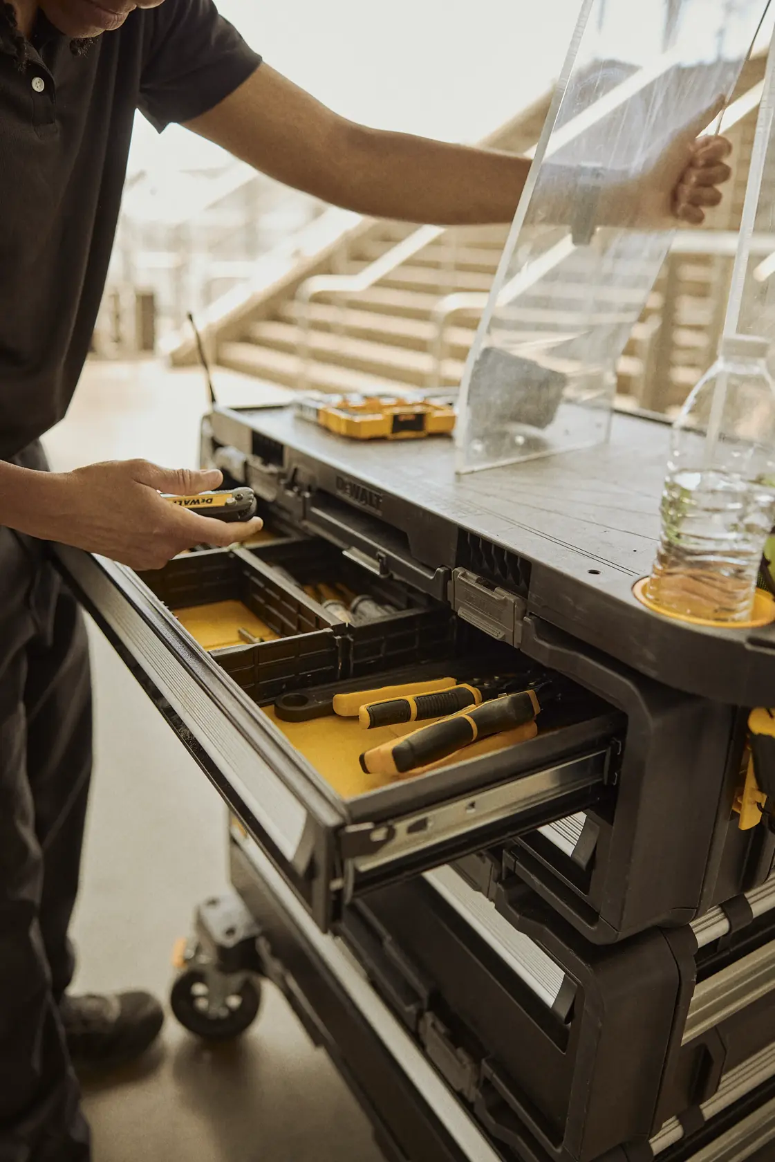A person accessing a drawer of a DEWALT mobile tool storage cabinet, which contains various hand tools and organized compartments. The cabinet is mounted on wheels for portability, and a transparent lid is being lifted. A water bottle is placed on top of the cabinet.