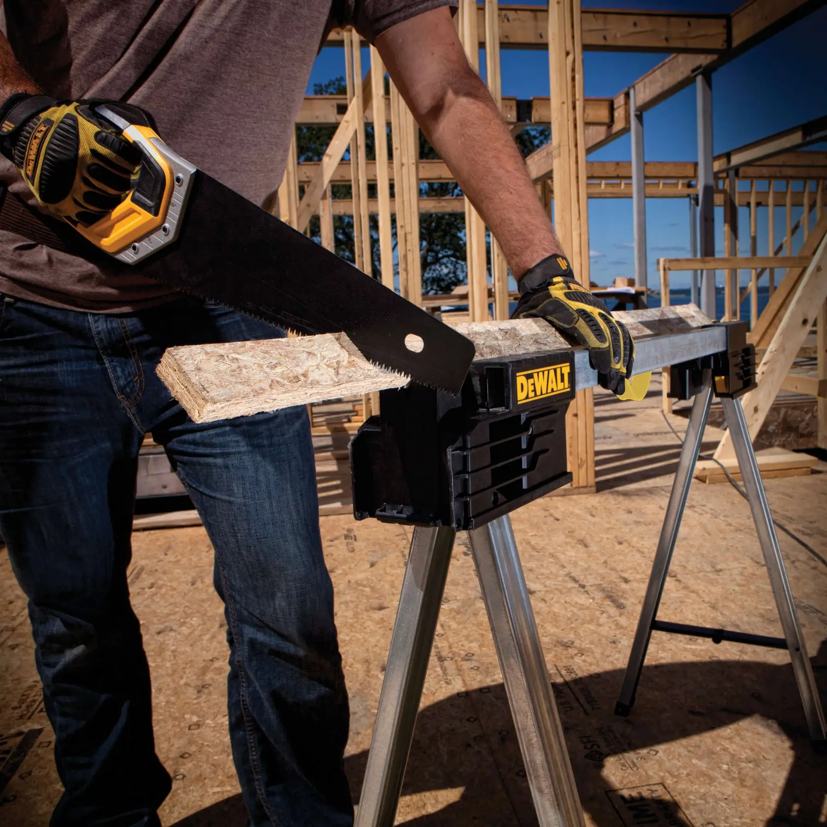 A person wearing work gloves uses a hand saw to cut a piece of wood resting on a DEWALT metal sawhorse at a construction site with wooden framing in the background.