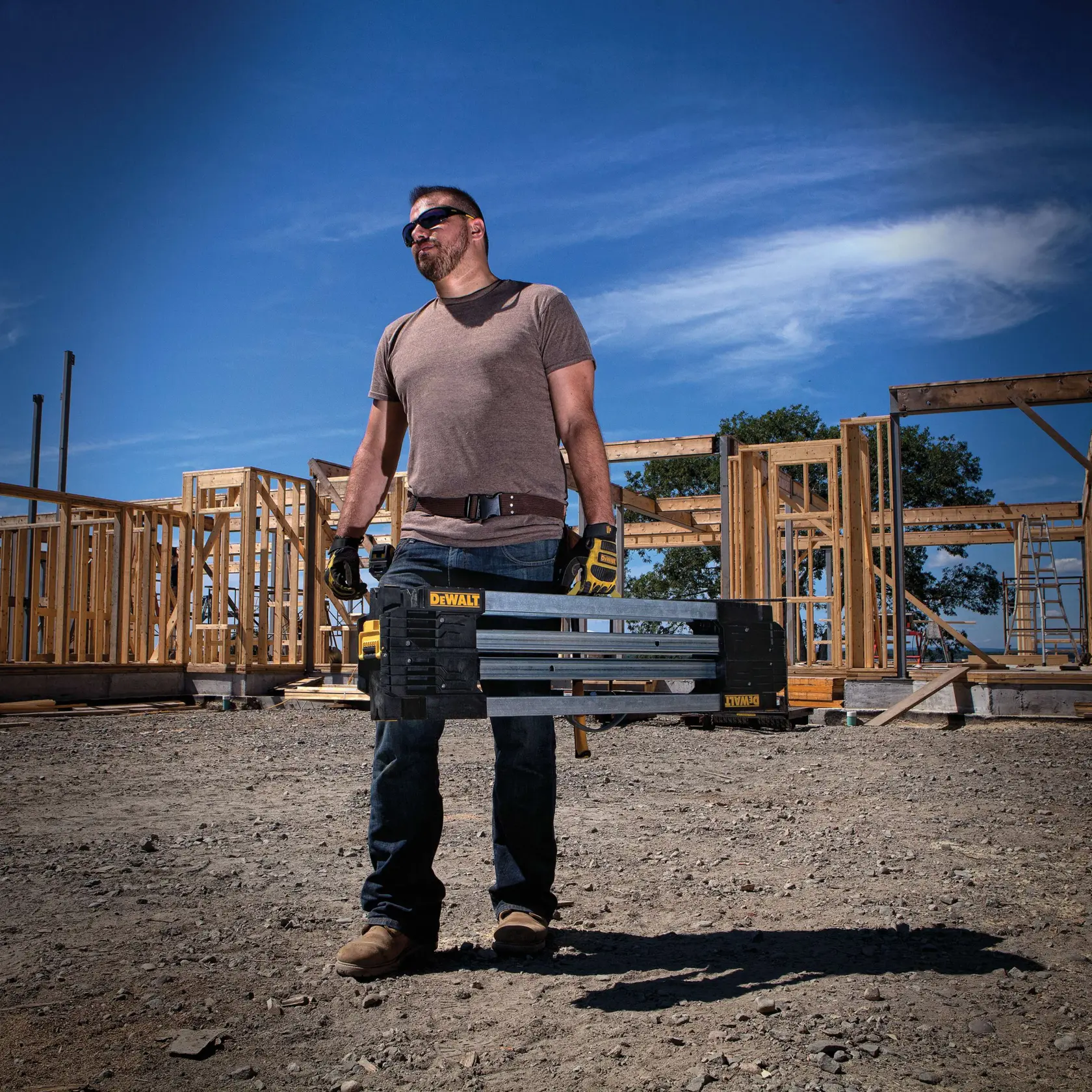 A person wearing gloves and work boots stands at a construction site, holding a DEWALT DWST11155 portable tool box. Wooden framing of buildings is visible in the background under a clear blue sky.
