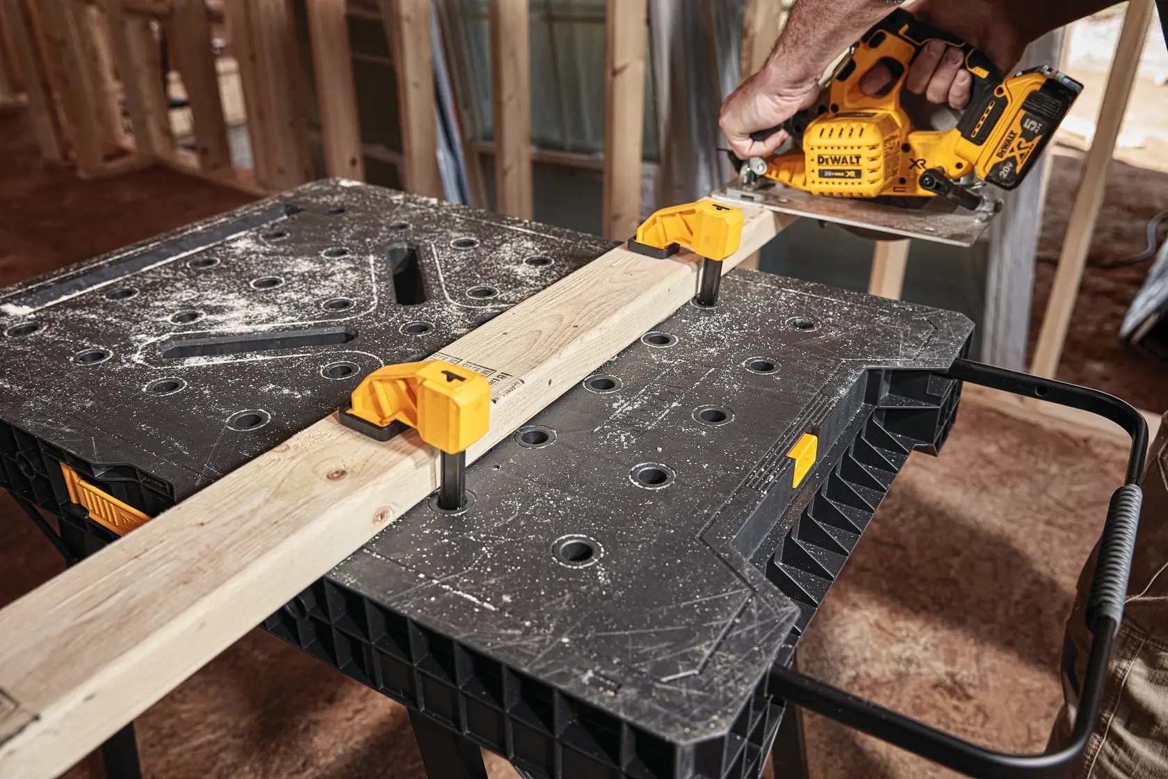 A DEWALT portable workbench with clamps securing a wooden plank, while a person uses a yellow DEWALT power tool to cut the wood in a construction setting.