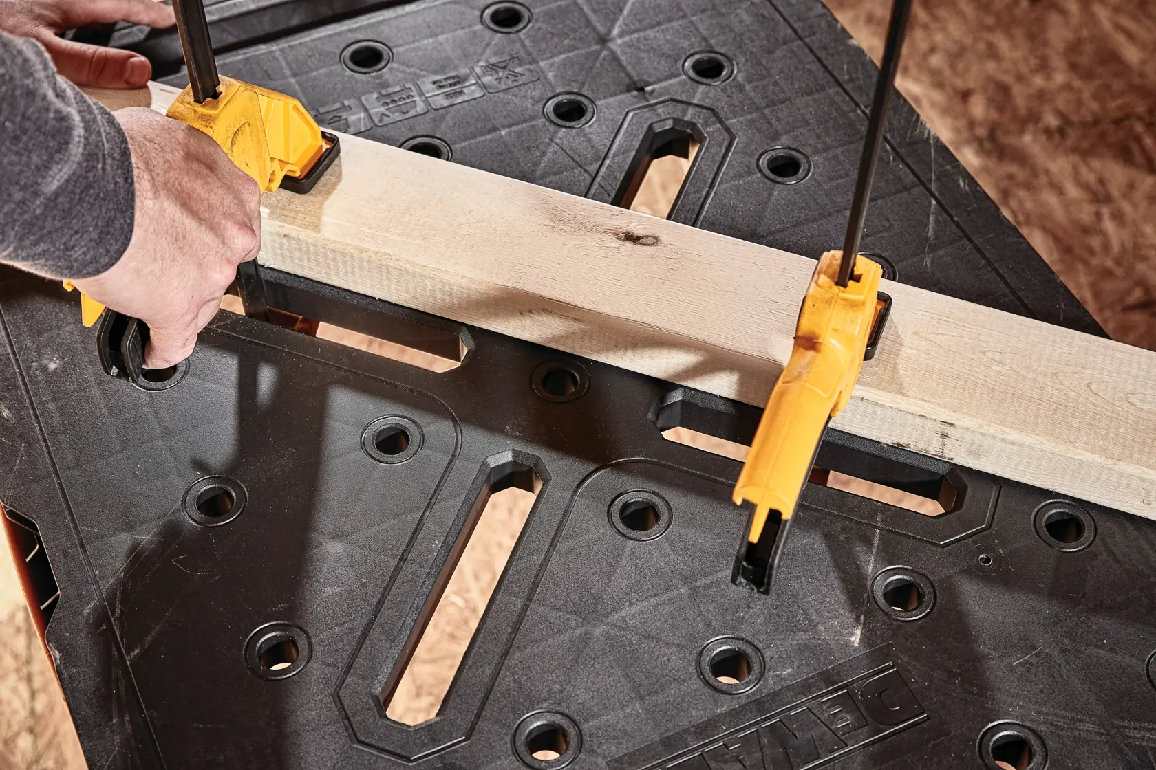 Close-up of a DEWALT workbench with a piece of wood clamped securely using two yellow clamps. A person's hands are adjusting one of the clamps. The black surface of the workbench has multiple holes and slots for versatility.
