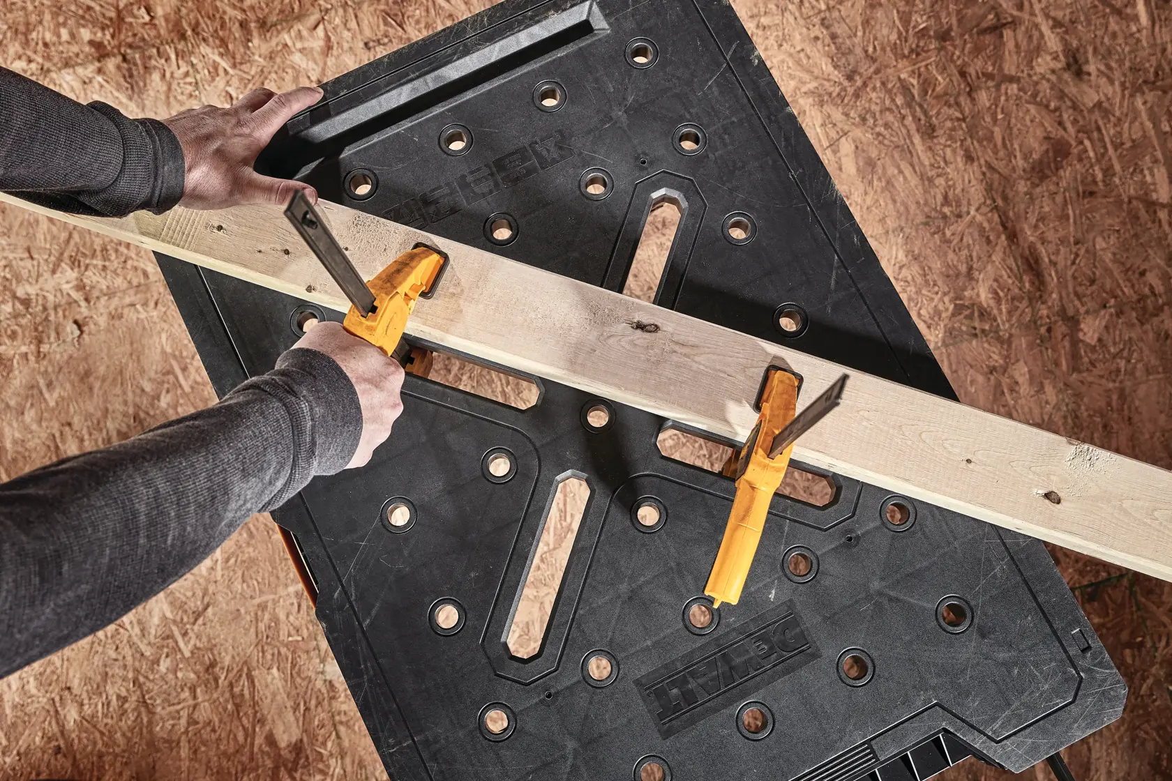 A person uses two yellow clamps to secure a wooden board onto a black DEWALT workbench.