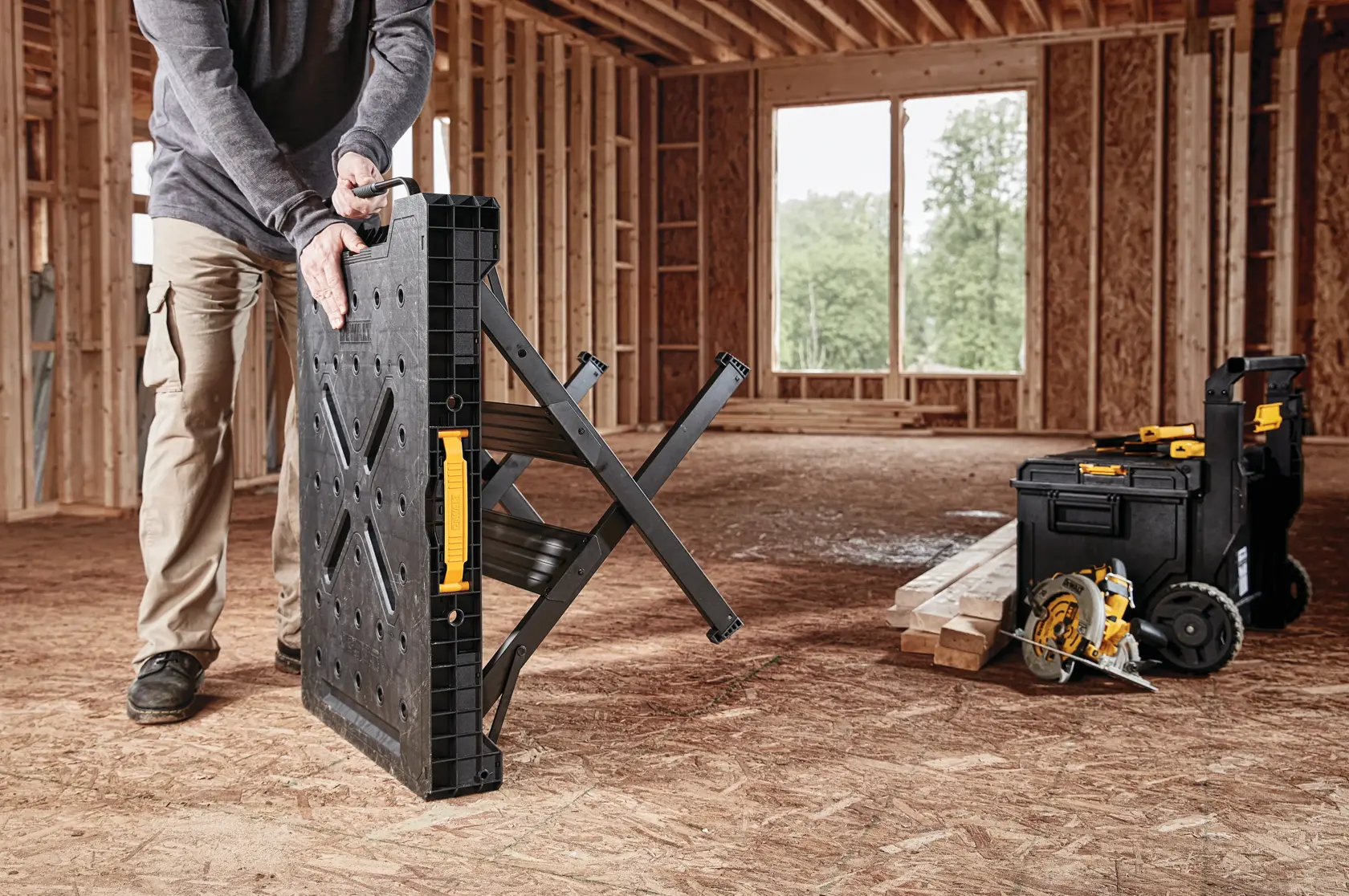 A person is unfolding a DEWALT portable workbench inside a building under construction. Next to the workbench are DEWALT toolboxes and a circular saw, placed on wooden boards.
