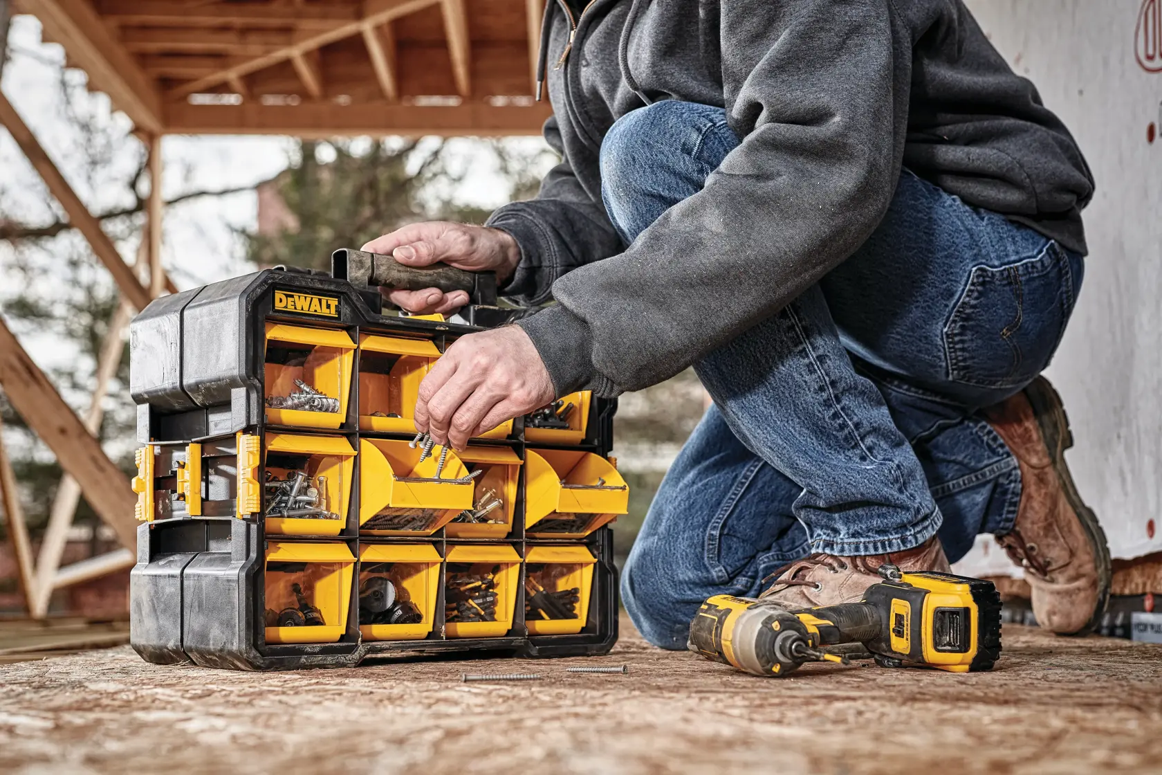A person is organizing hardware items in a DEWALT portable storage organizer with yellow compartments. The organizer is on the ground outdoors, and a DEWALT power drill lies next to it.