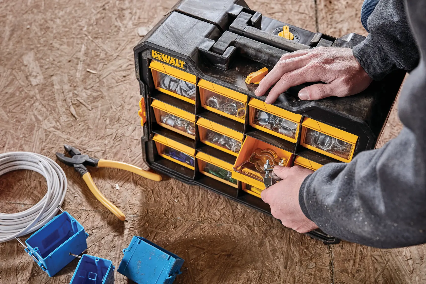 A person is organizing small hardware parts in a DEWALT storage organizer with multiple compartments. The organizer is placed on a wooden surface, beside a pair of pliers, electrical wire, and blue plastic outlet boxes.