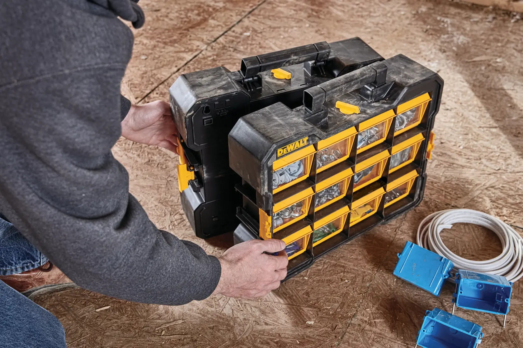 A person handles a DEWALT DWST14121 portable organizer with yellow compartments and a black case, filled with hardware and small parts, placed on a wooden floor next to electrical supplies.
