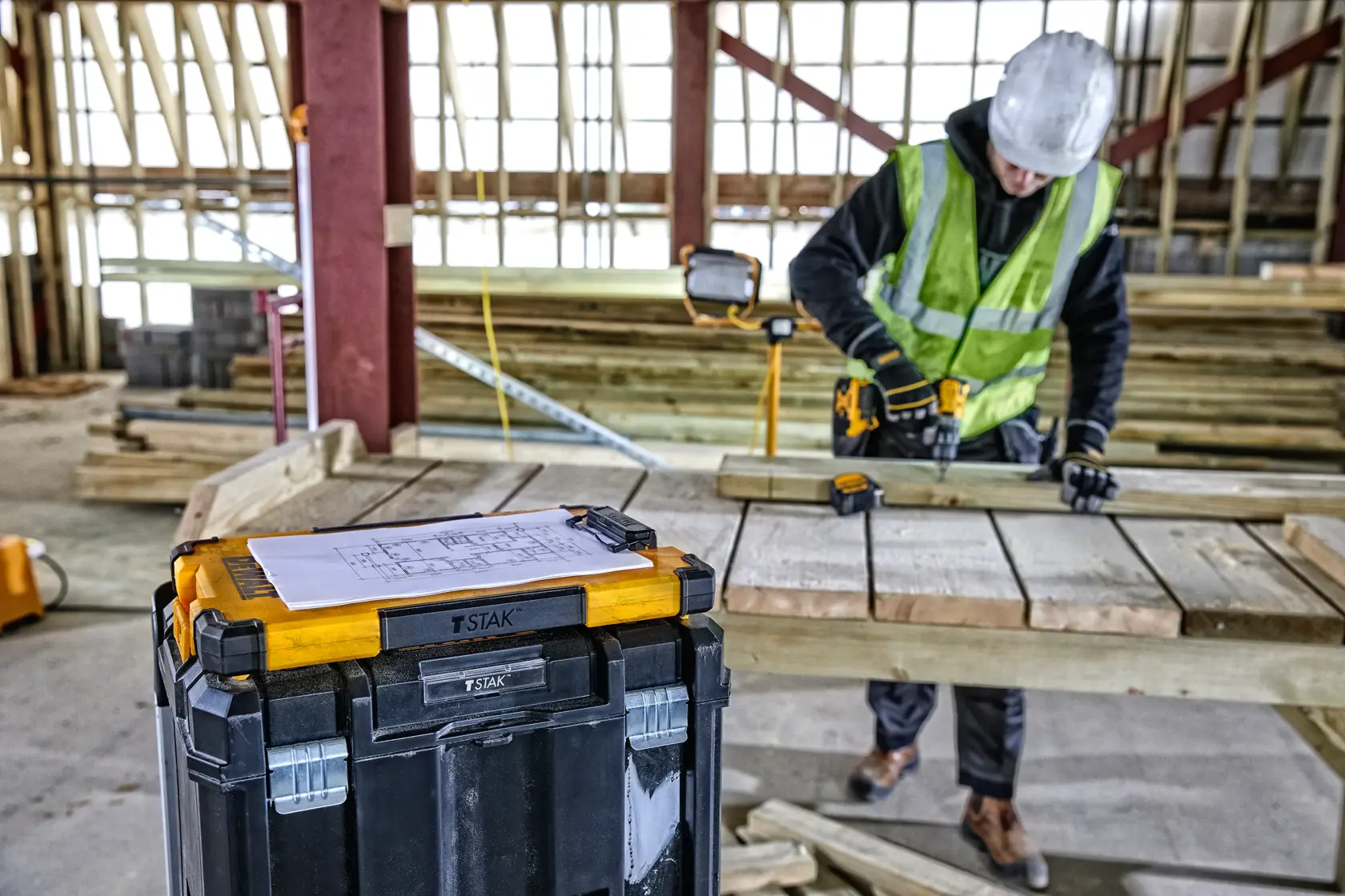 A DEWALT TSTAK tool storage box with a paper on top is positioned in the foreground, while a construction worker wearing a safety vest and helmet uses a power drill on wooden planks in the background of a construction site.